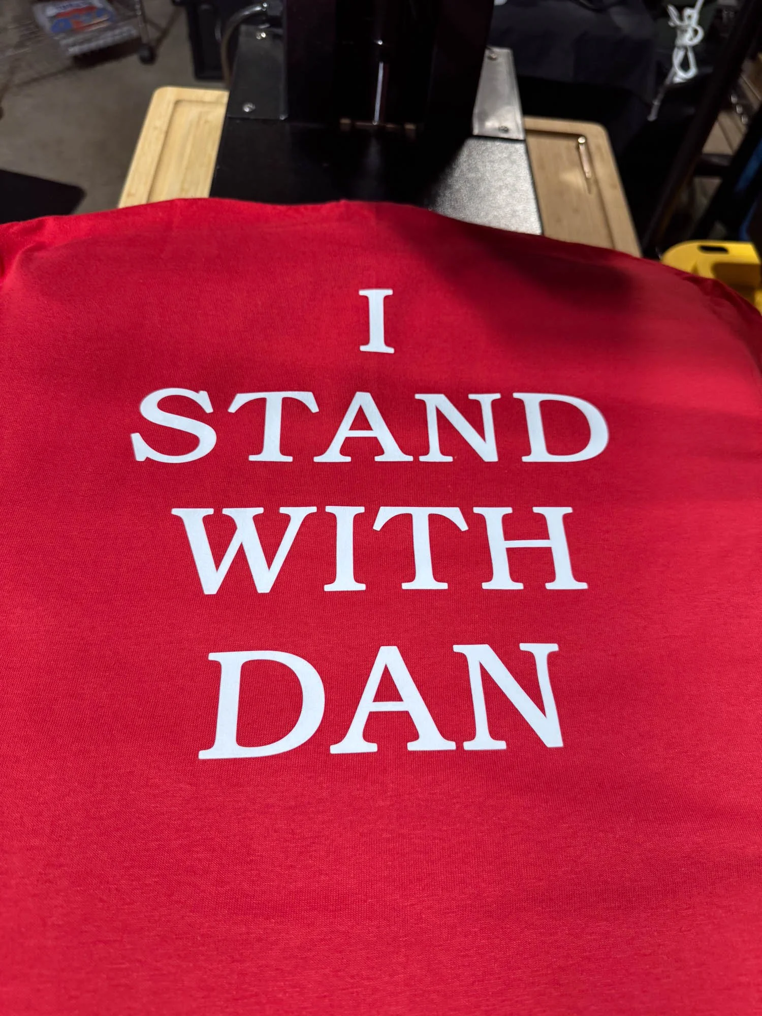 Red shirt with white text that reads 'I STAND WITH DAN' lying on a flat surface.