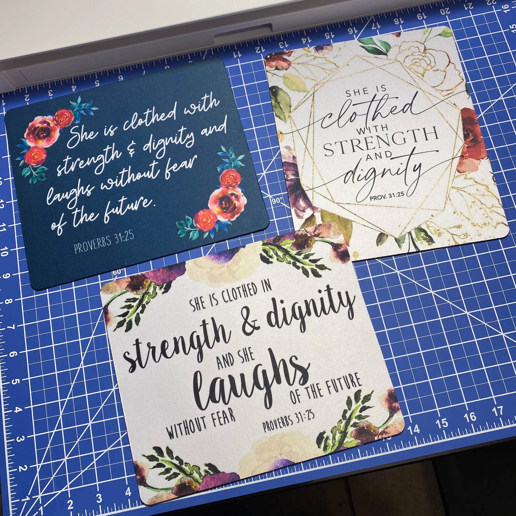 Three decorative cards with inspirational Bible quotes on a blue cutting mat. The quotes are from Proverbs 31:25 and feature floral designs and elegant fonts.