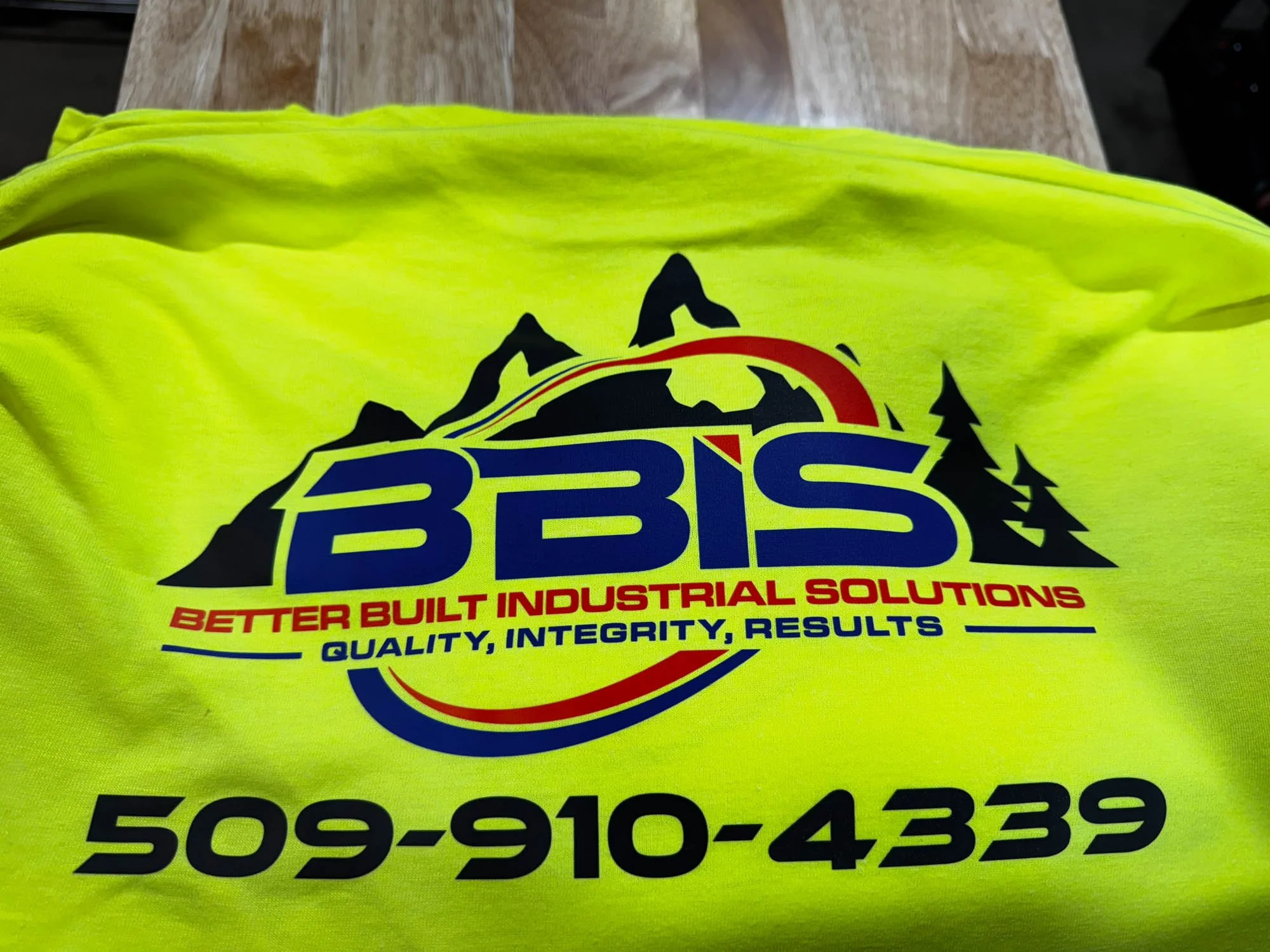 Bright yellow shirt with logo and text for BBS Better Built Industrial Solutions. Logo features mountains, trees, and a semi-circle in red, yellow, and blue. Phone number 509-910-4339.