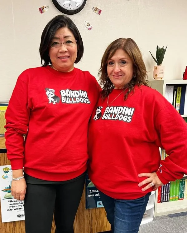 Two women wearing red 'Bandini Bulldogs' sweatshirts pose in front of a bookshelf and a wall with a clock and decorative items, smiling at the camera.