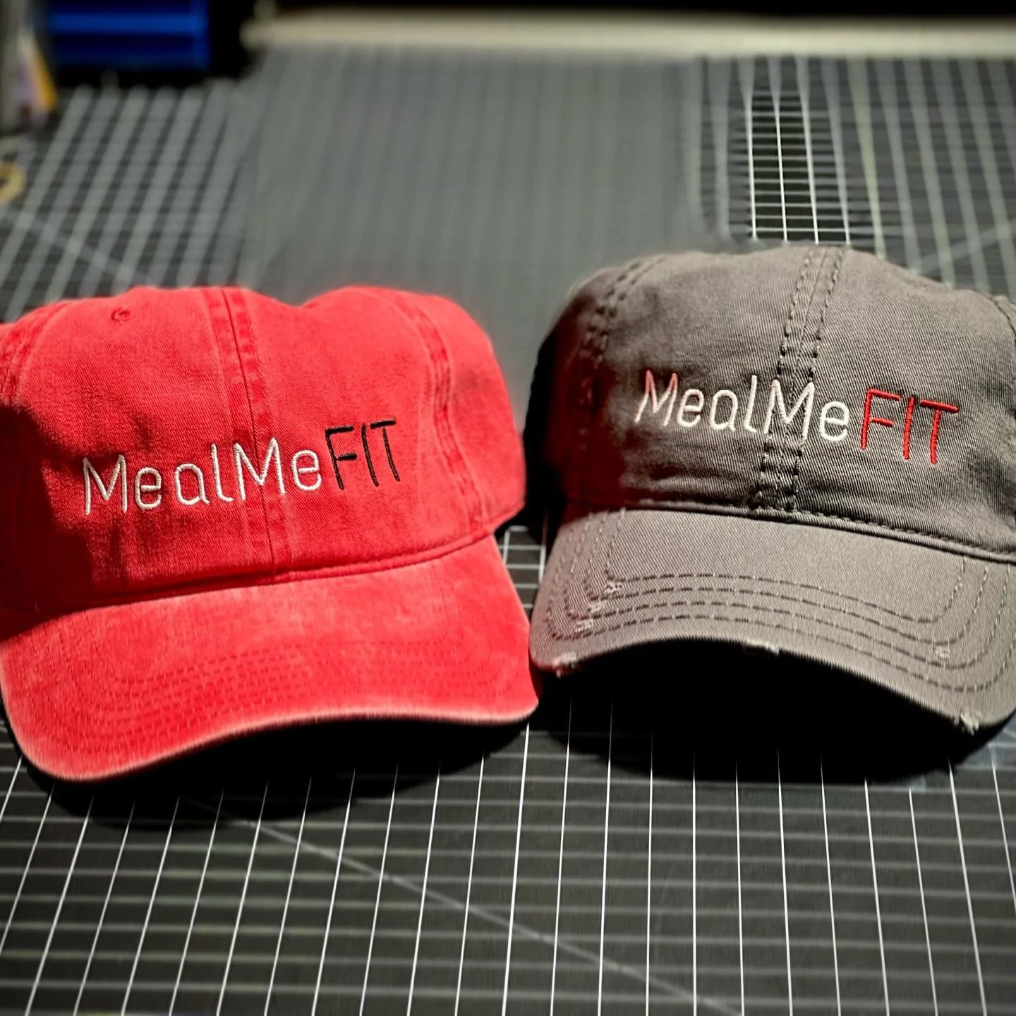 Two baseball caps, one red and one gray, with embroidered text 'MealMeFIT'; the red cap has the text in white and black, the gray cap has white and red letters, placed on a black surface with a grid pattern.