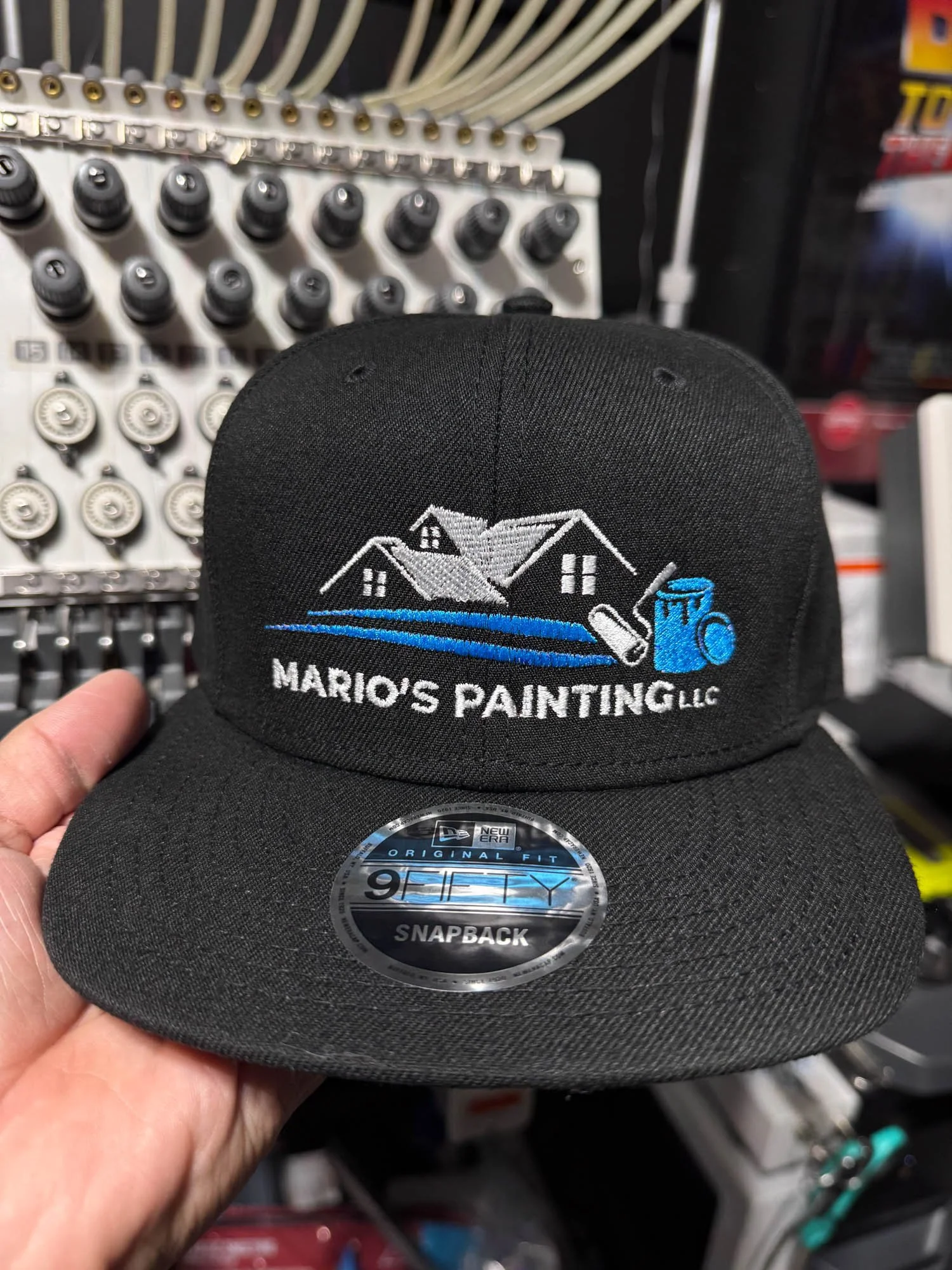 Black baseball cap with embroidered logo of houses, a roller paintbrush, and a paint can, with the text "Mario's Painting LLC," held in front of a display of spray paint cans.