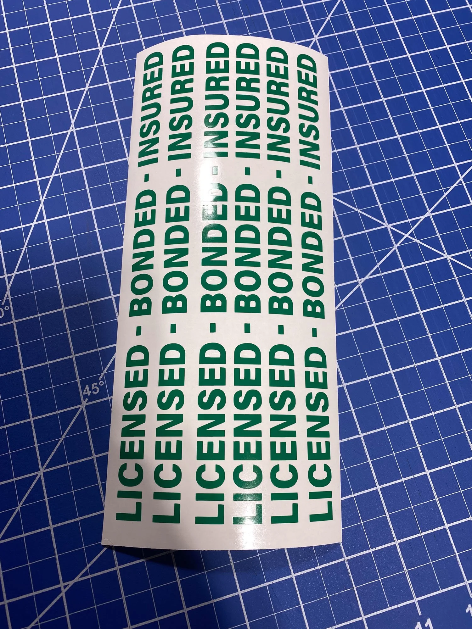 Sheet of license stickers with green text reading "LICENSED - BONDED - INSURED" on a blue cutting mat.