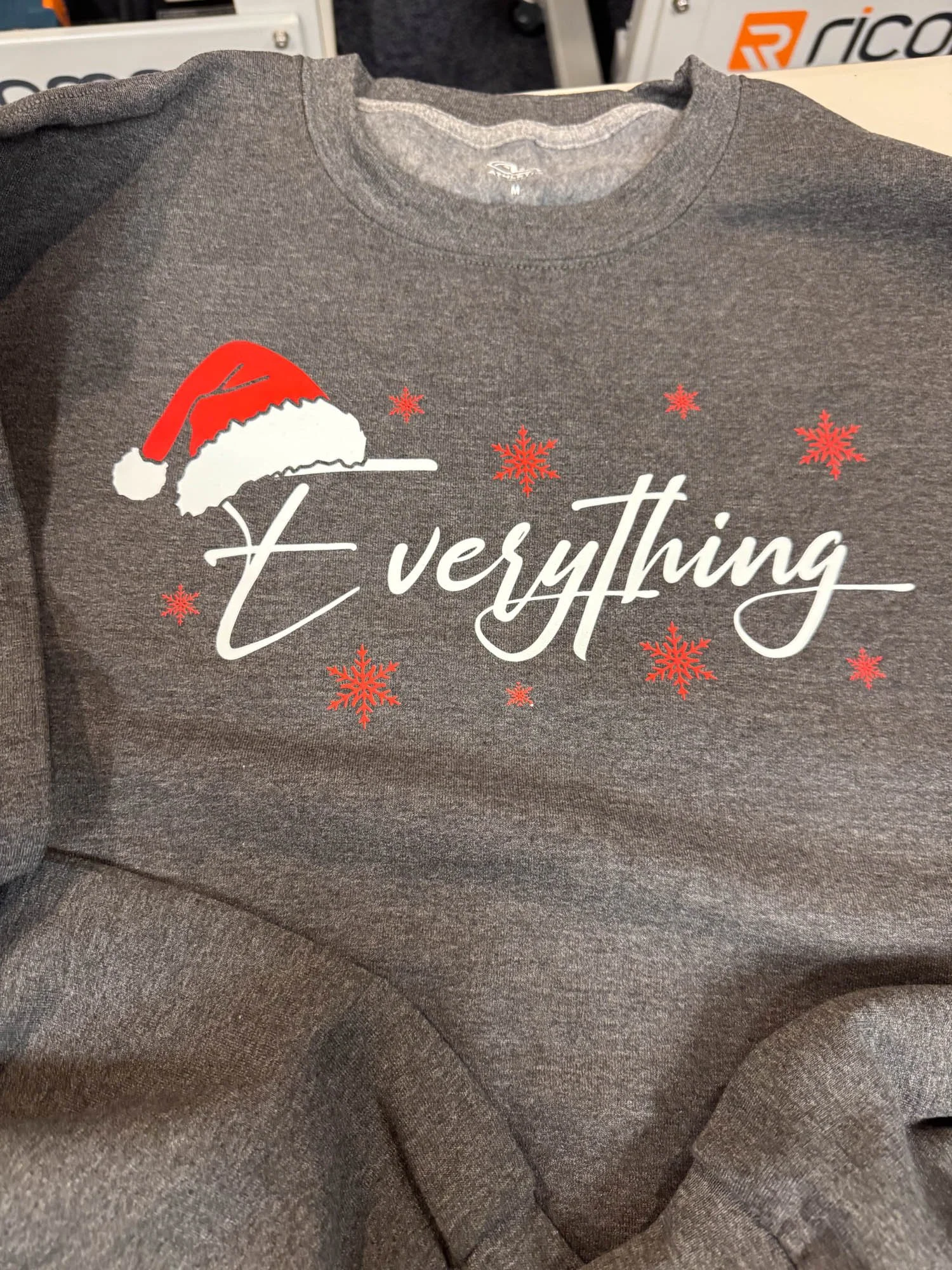 A gray sweatshirt with a Christmas theme, featuring a Santa hat and red snowflakes, and the partial phrase 't verything' printed on it.