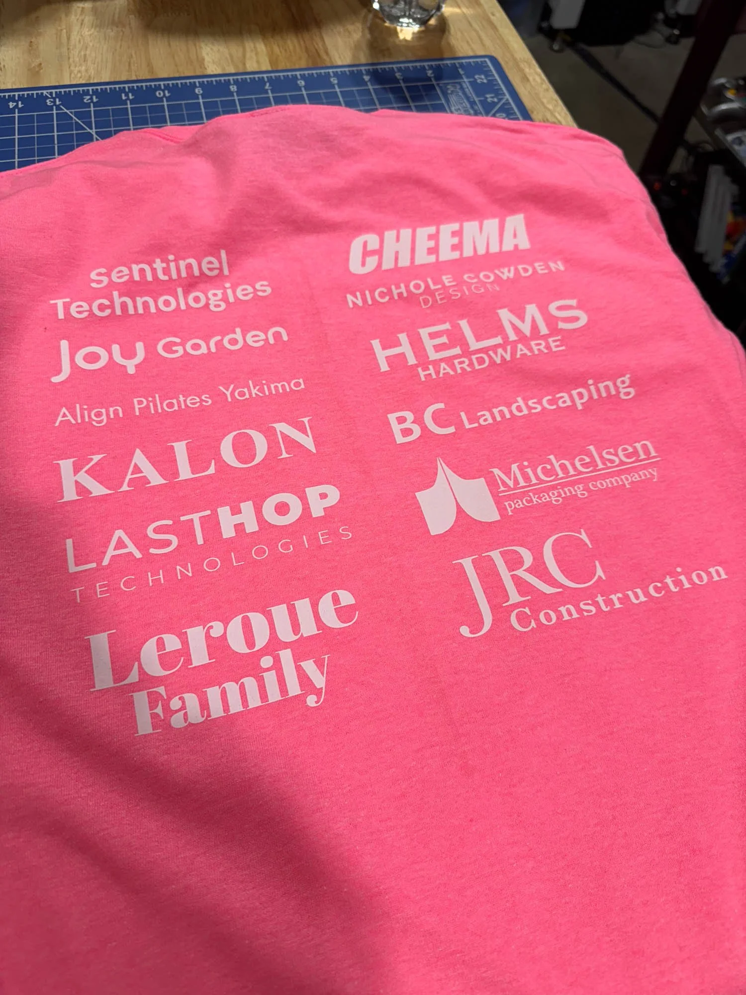 Pink T-shirt with various company logos and names printed in white, laid on a cutting mat on a wooden table.