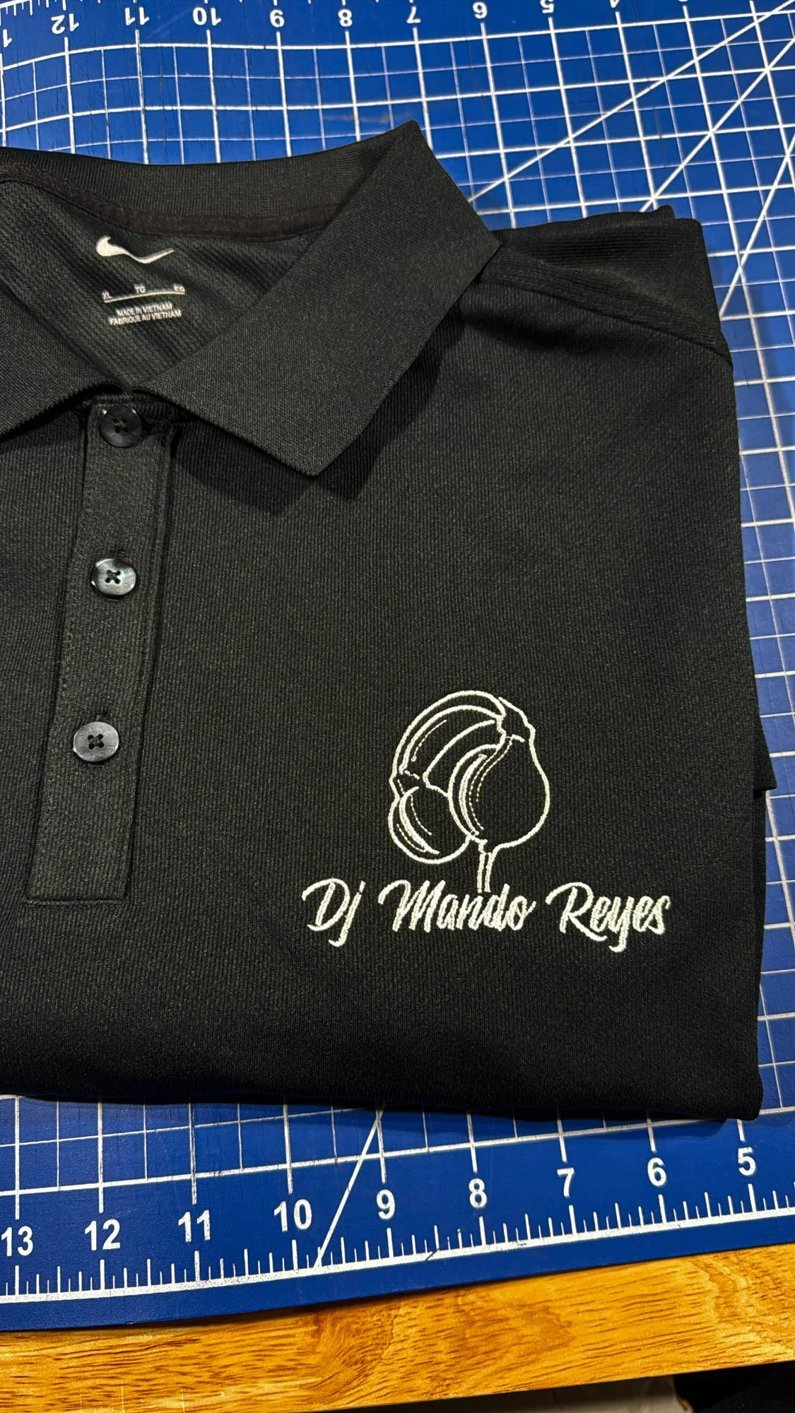 Black polo shirt with white embroidered logo of headphones and text 'DJ Mando Reyes' on a blue cutting mat.