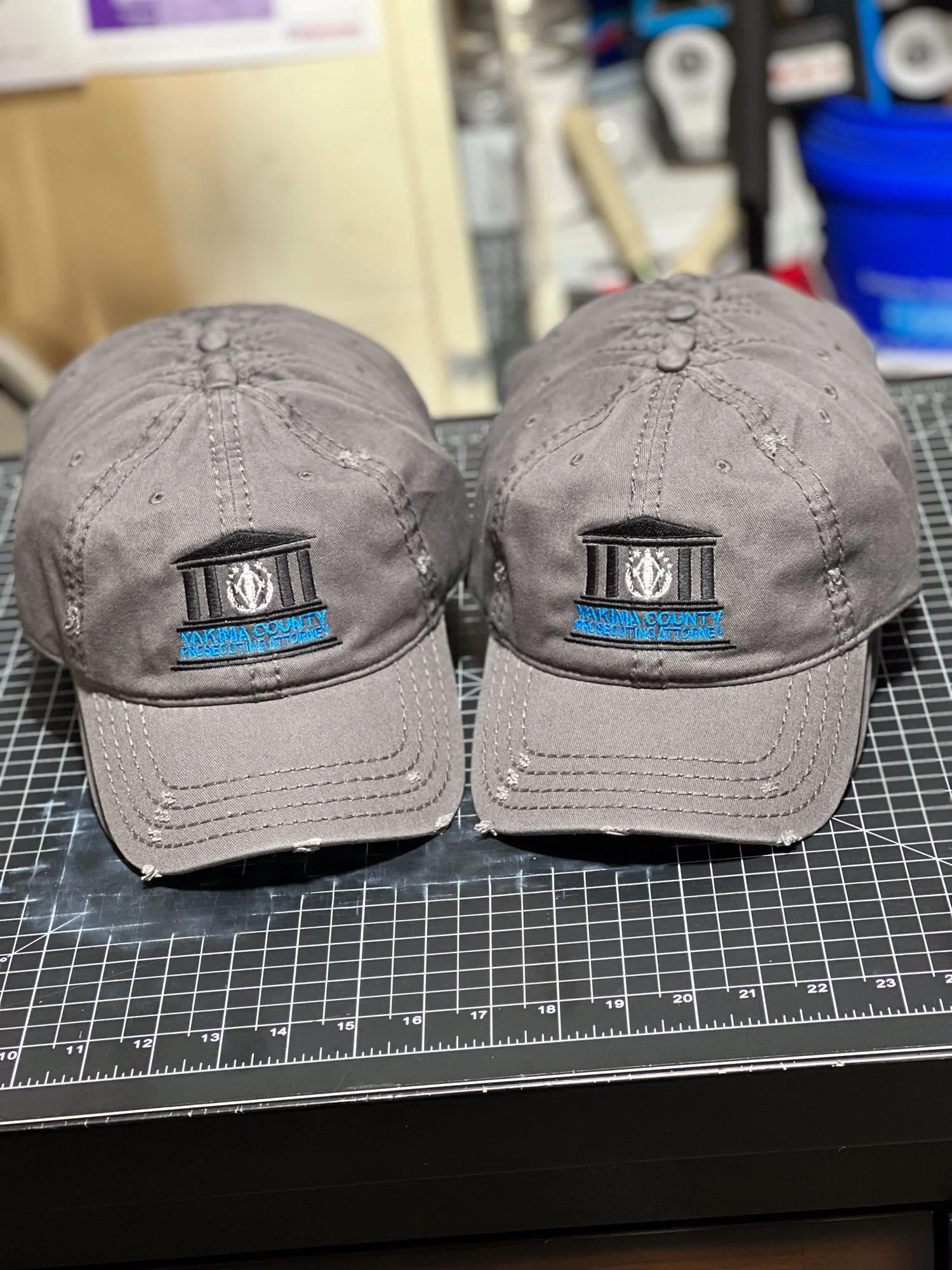 Two gray caps embroidered with the logo of the Yakima County Prosecuting Attorney, placed on a black grid cutting mat.