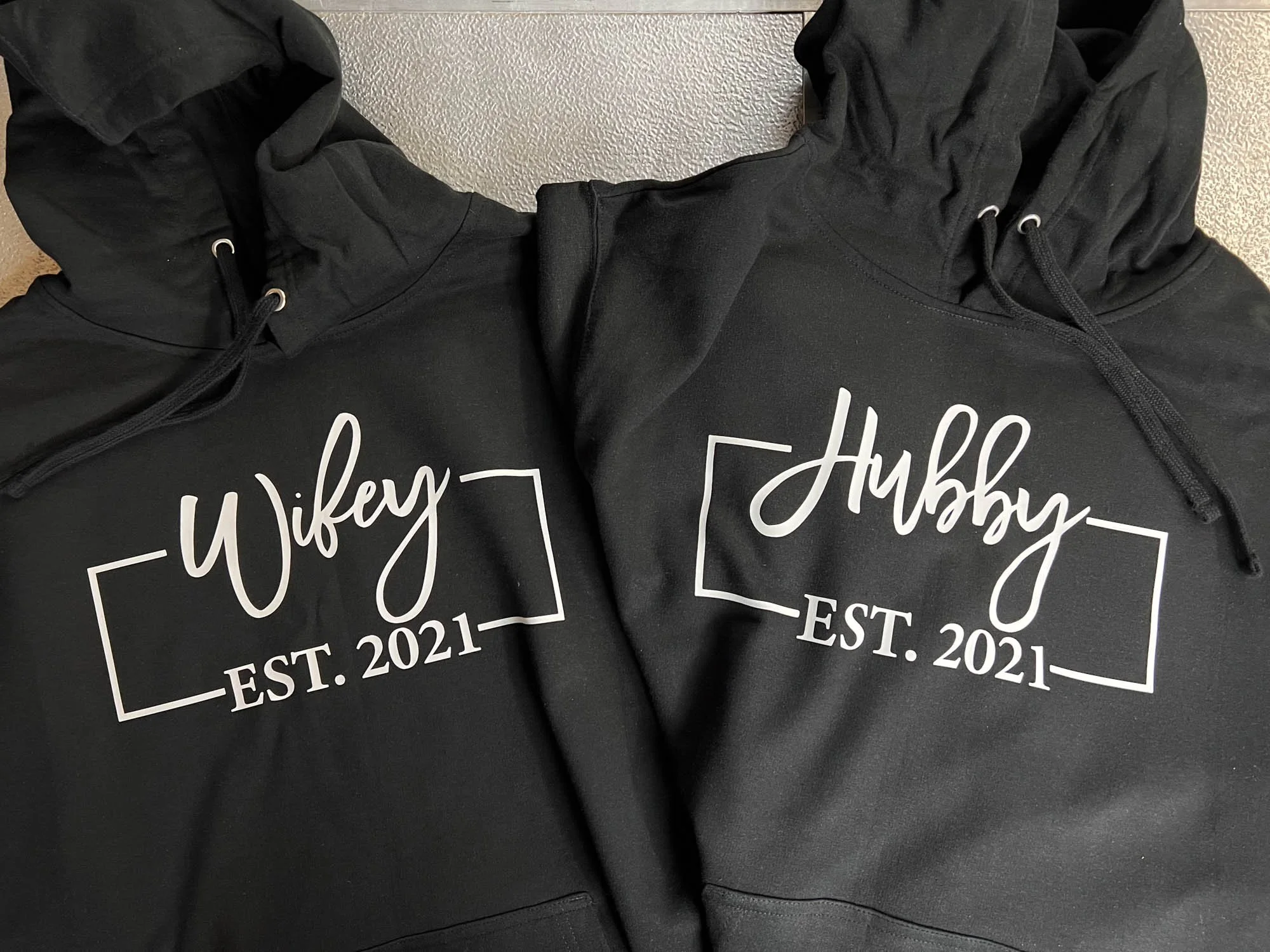 Two black hoodies with white text reading "Wifey" and "Hubby," each with an "EST. 2021" underneath, laid flat on a surface.