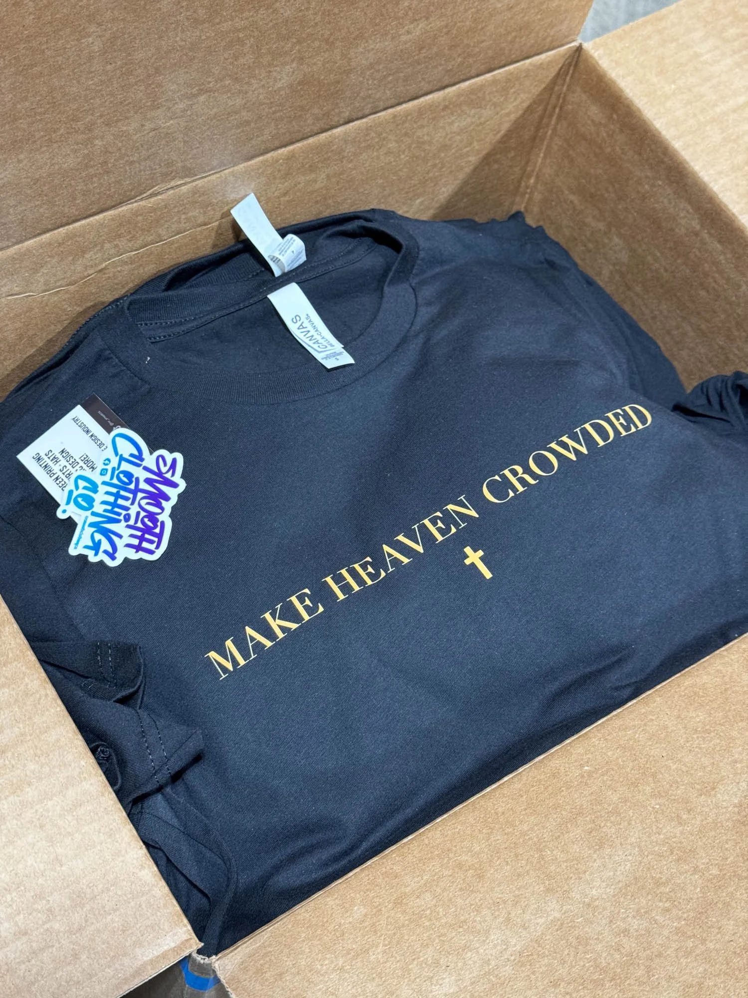 Black T-shirt inside a cardboard box with the text "Make Heaven Crowded" and a plus sign, along with some tags attached.