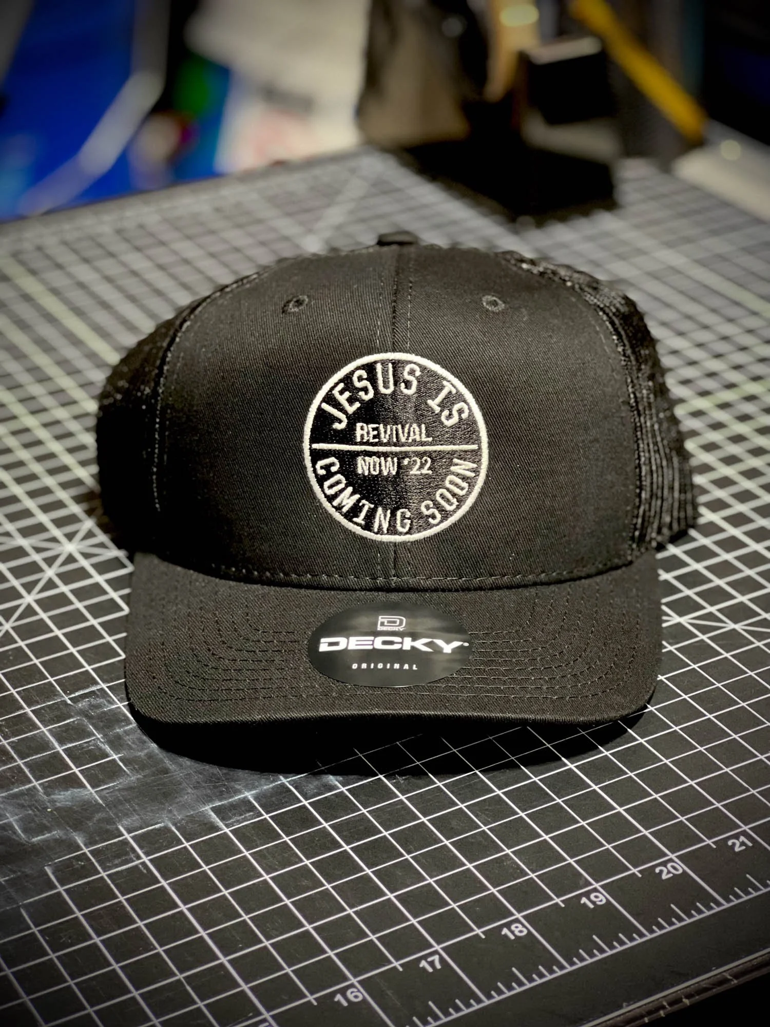 A black baseball cap with white embroidery that says 'JESUS T'S COMING' and 'REVIVAL NOV '22'. The cap is placed on a black cutting mat with a grid pattern.