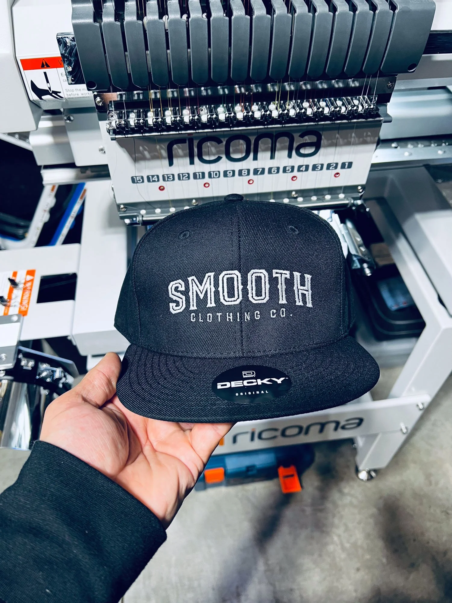 A person holding a black cap with 'SMOOTH CLOTHING CO.' embroidered on the front, in front of a Ricoma embroidery machine.