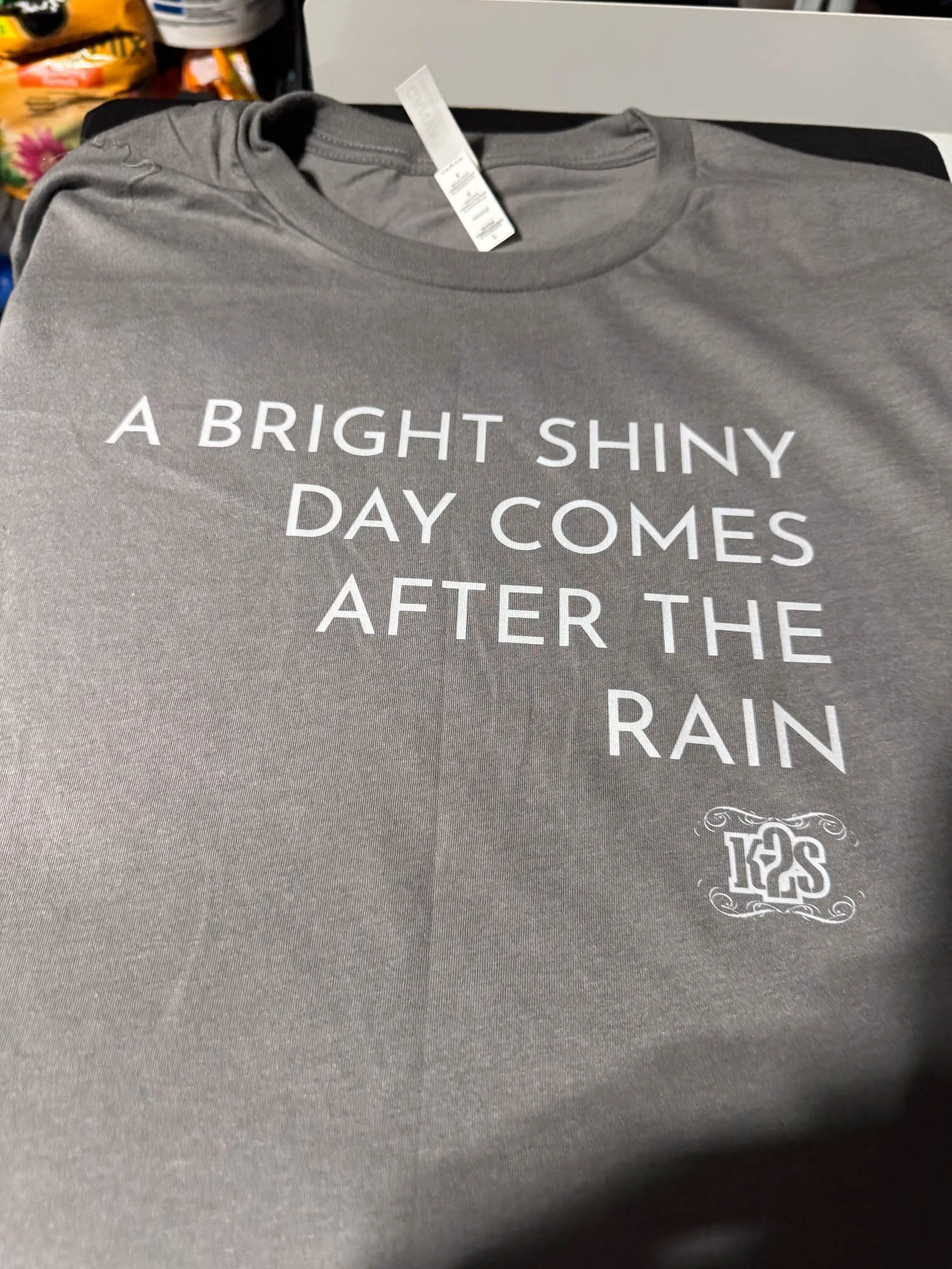 Gray T-shirt with white text that reads, 'A BRIGHT SHINY DAY COMES AFTER THE RAIN,' and a small logo with the text 'K2S' at the bottom right.