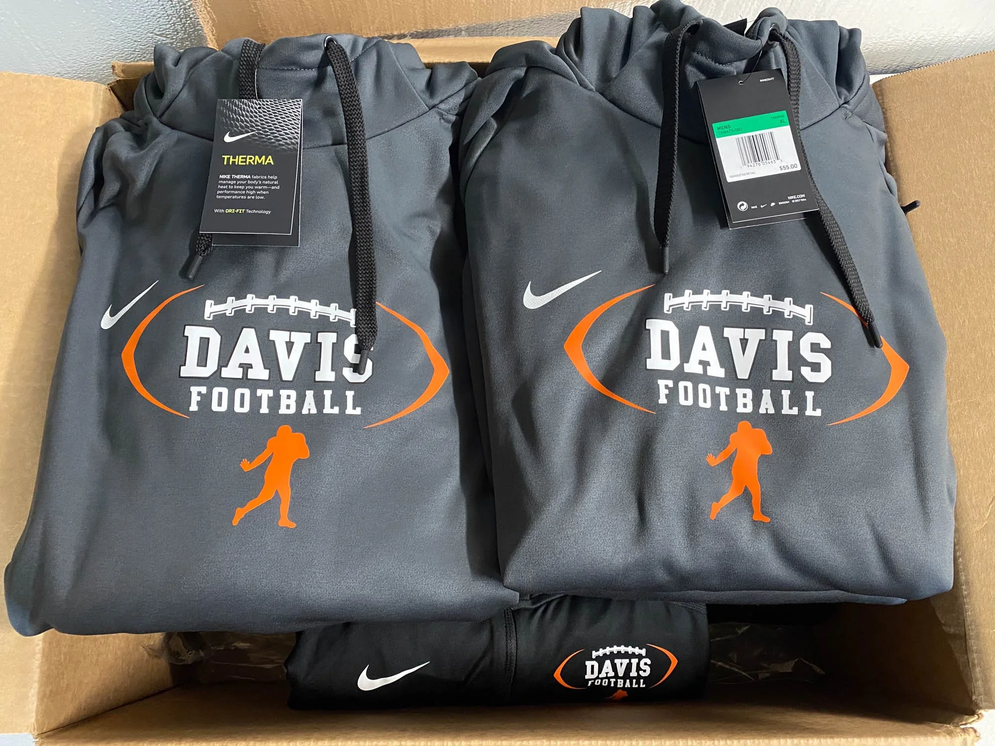 Two gray Nike football jackets with 'Davis Football' logo and orange football graphics, placed inside a cardboard box. One jacket has tags, one is priced at $55, and a black Nike bag with logo is also inside.