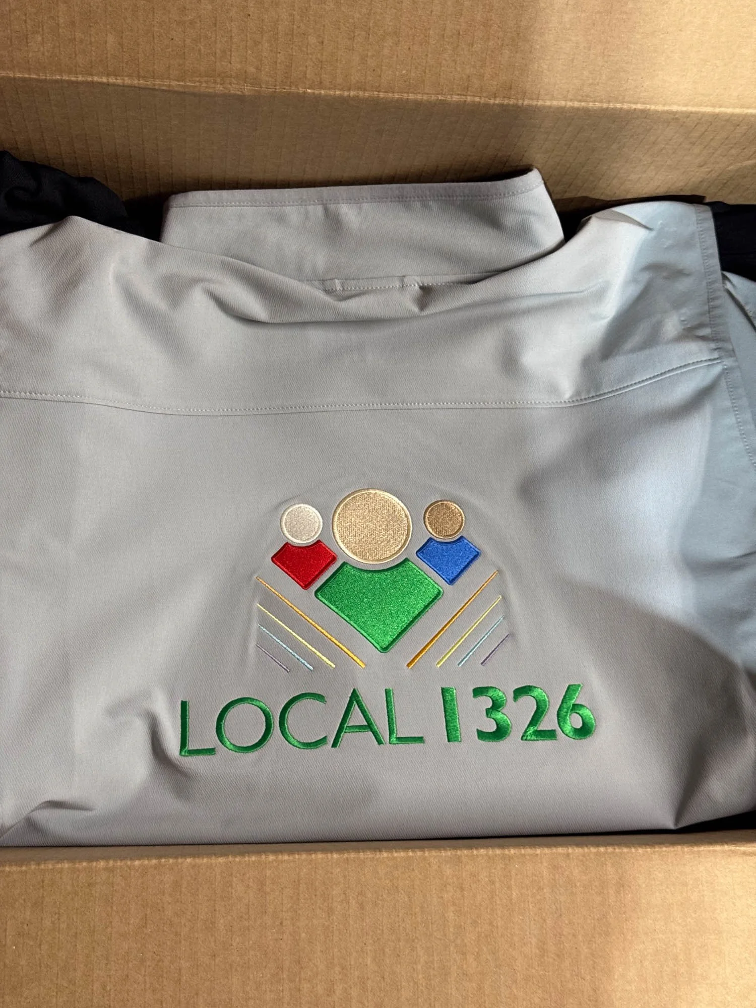 A gray shirt with embroidered text 'LOCAL 1326' and a logo of three stylized human figures in red, gold, and blue above the text, inside a cardboard box.