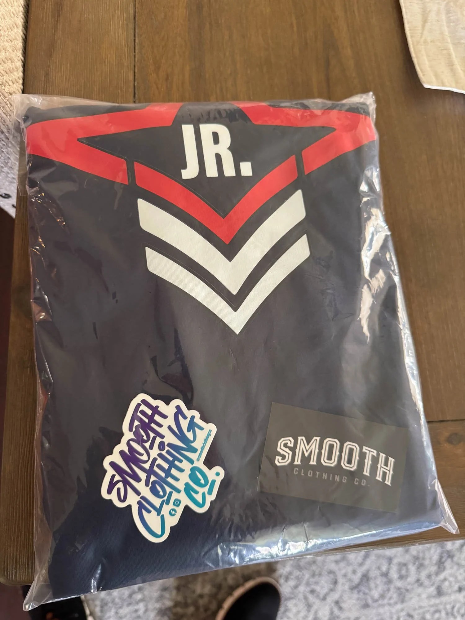 A folded black shirt in plastic packaging with large white chevron designs, a red star, and the word 'JR.' on top. It has two stickers, one with colorful text reading 'Smoother Clothing Co.!' and the other with the brand name 'Smooth Clothing Co.'