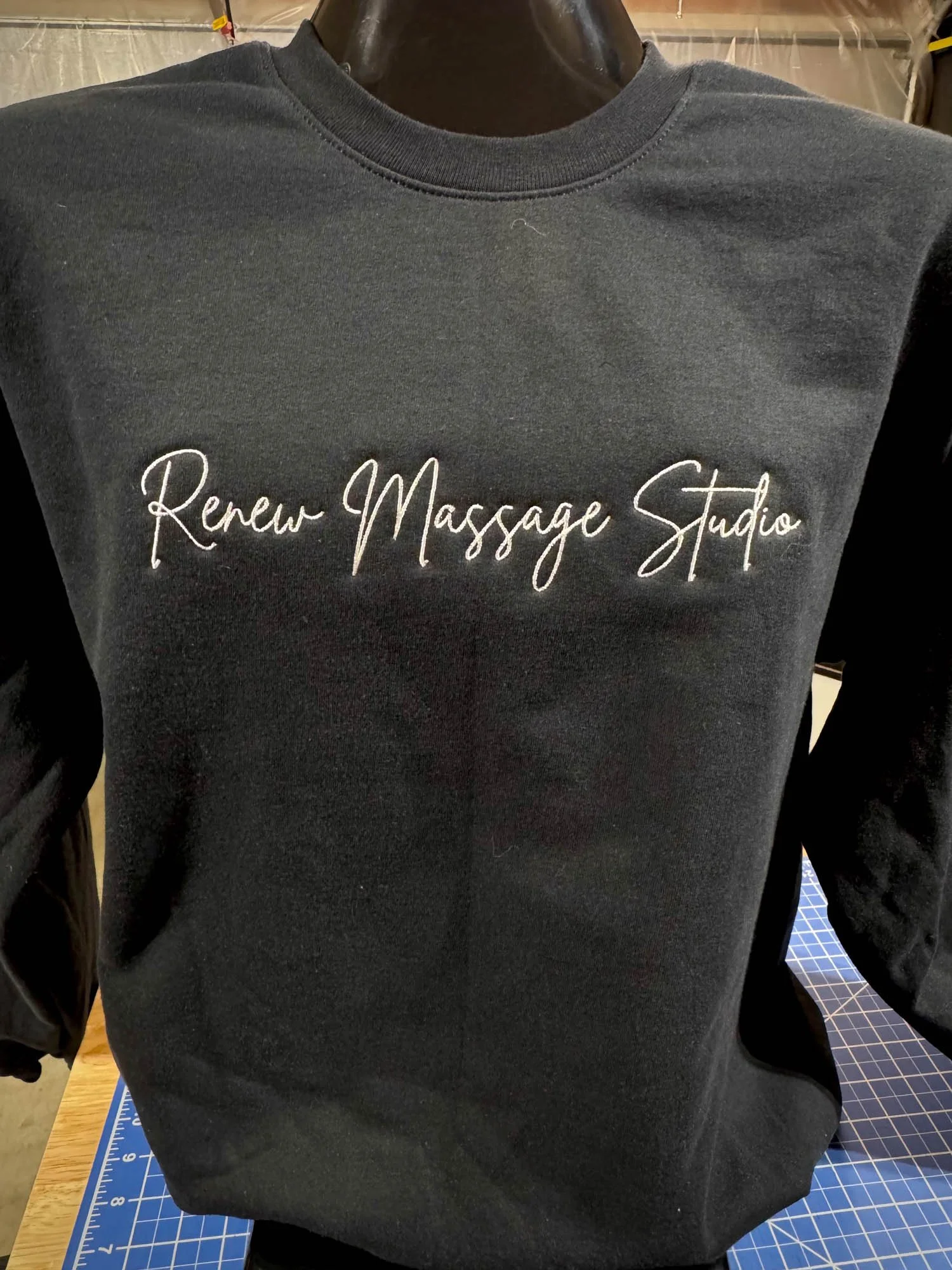 Black sweatshirt with white script text that reads 'Renew Massage Studio'. Mennequin torso, background of a work table and industrial setting.