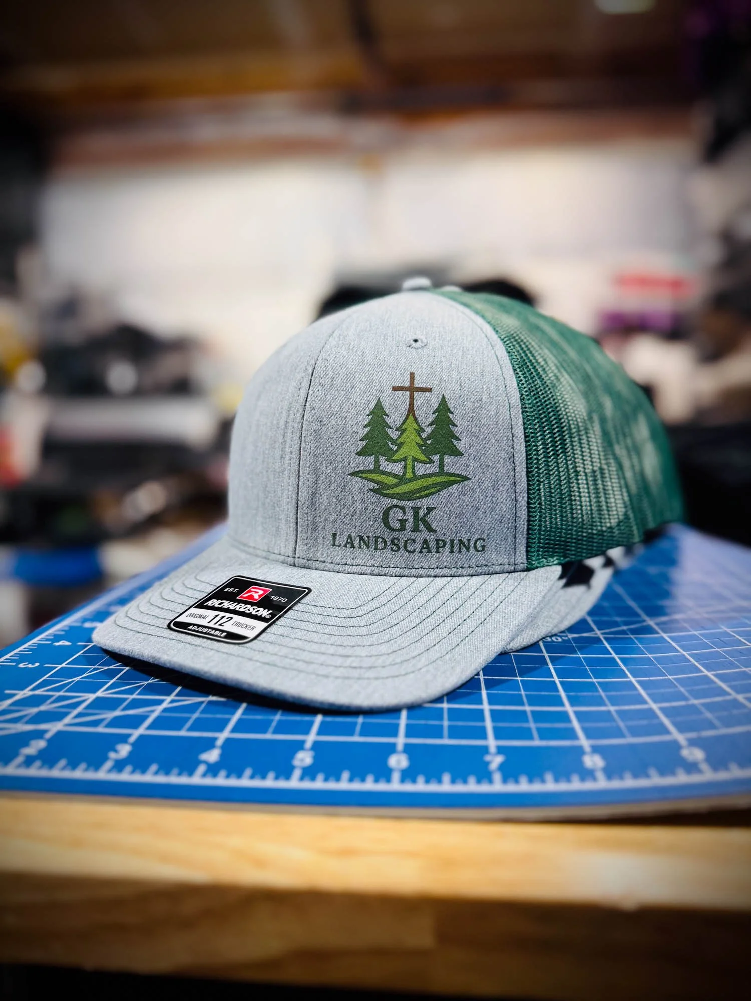 Gray and green baseball cap with the logo of GK Landscaping, featuring three evergreen trees and a cross. The cap is placed on a blue cutting mat with grid lines. The background appears to be cluttered or a workshop.