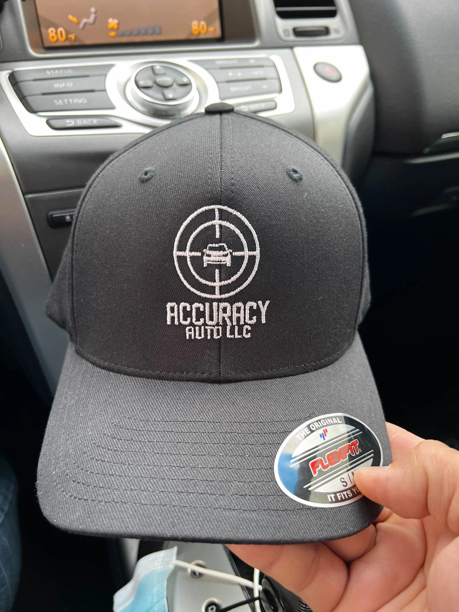 A gray and black baseball cap with embroidered logo of a car in crosshairs and the words 'ACCURACY AUTO LLC' being held inside a car near the center console.