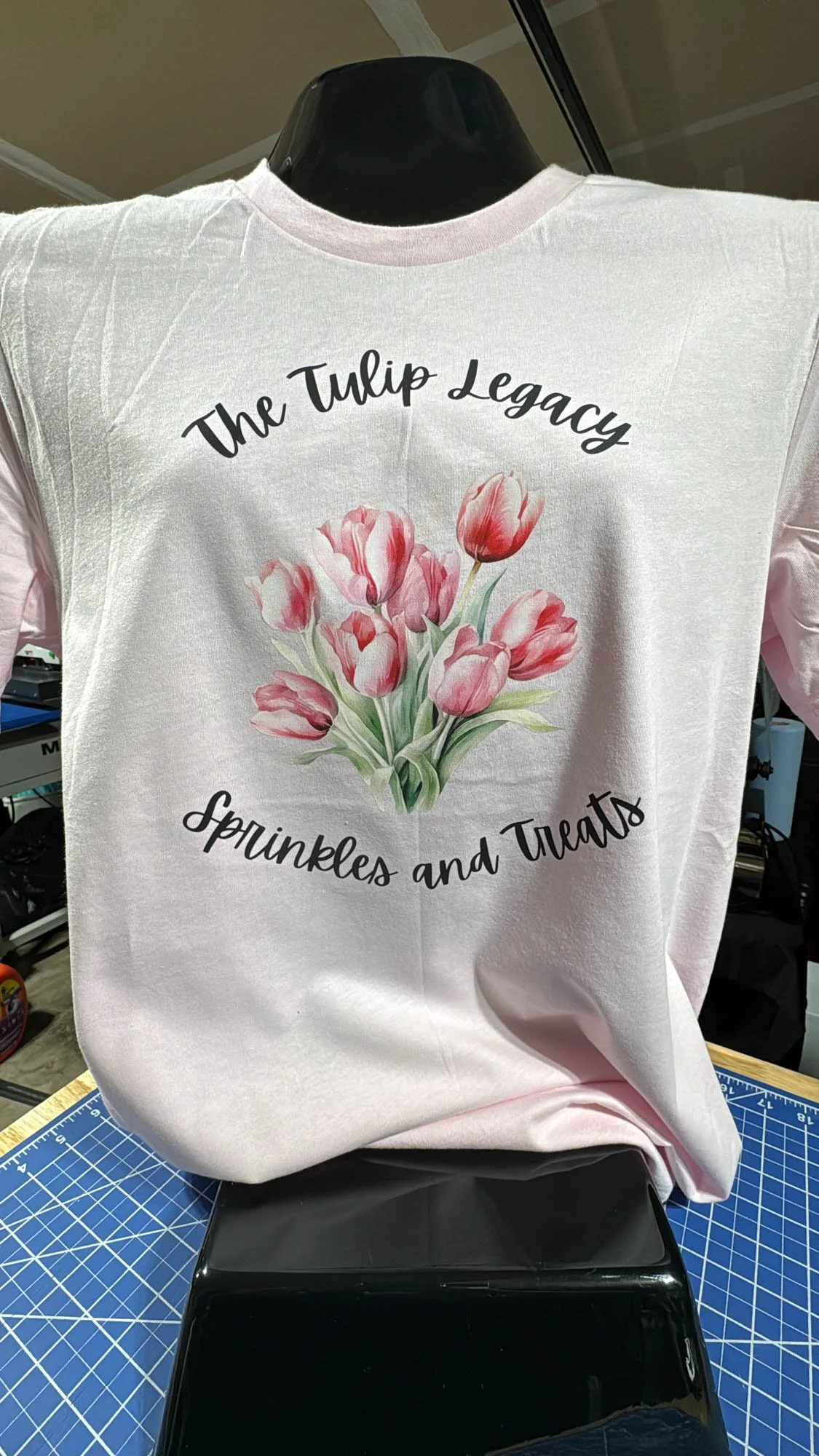 A white t-shirt with a pink and green tulip bouquet graphic and the text 'The Tulip Legacy' above and 'Sprinkles and Treats' below, displayed on a black mannequin torso.