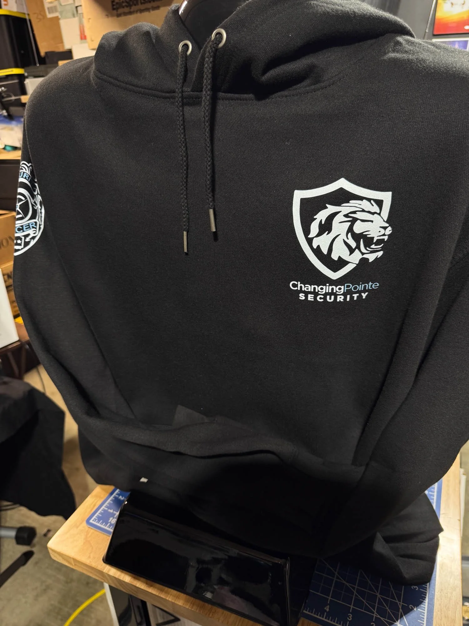 A black hoodie with a logo of a lion's head inside a shield and the text 'Changing Pointe Security' on the chest, displayed on a mannequin or stand on a workbench in a workshop or store.