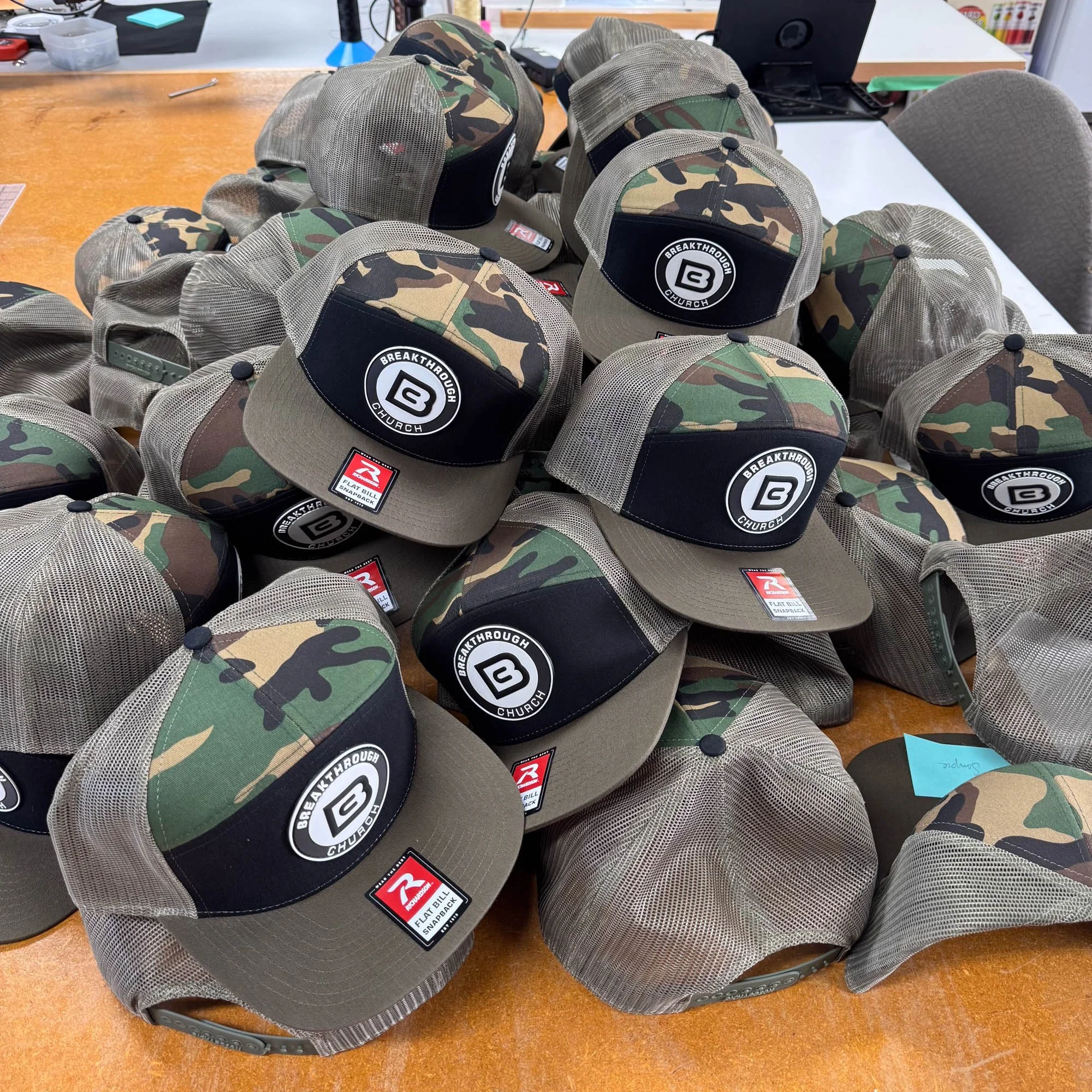 Multiple camouflage and beige trucker hats with 'Breakthrough Church' logo on a wooden table.