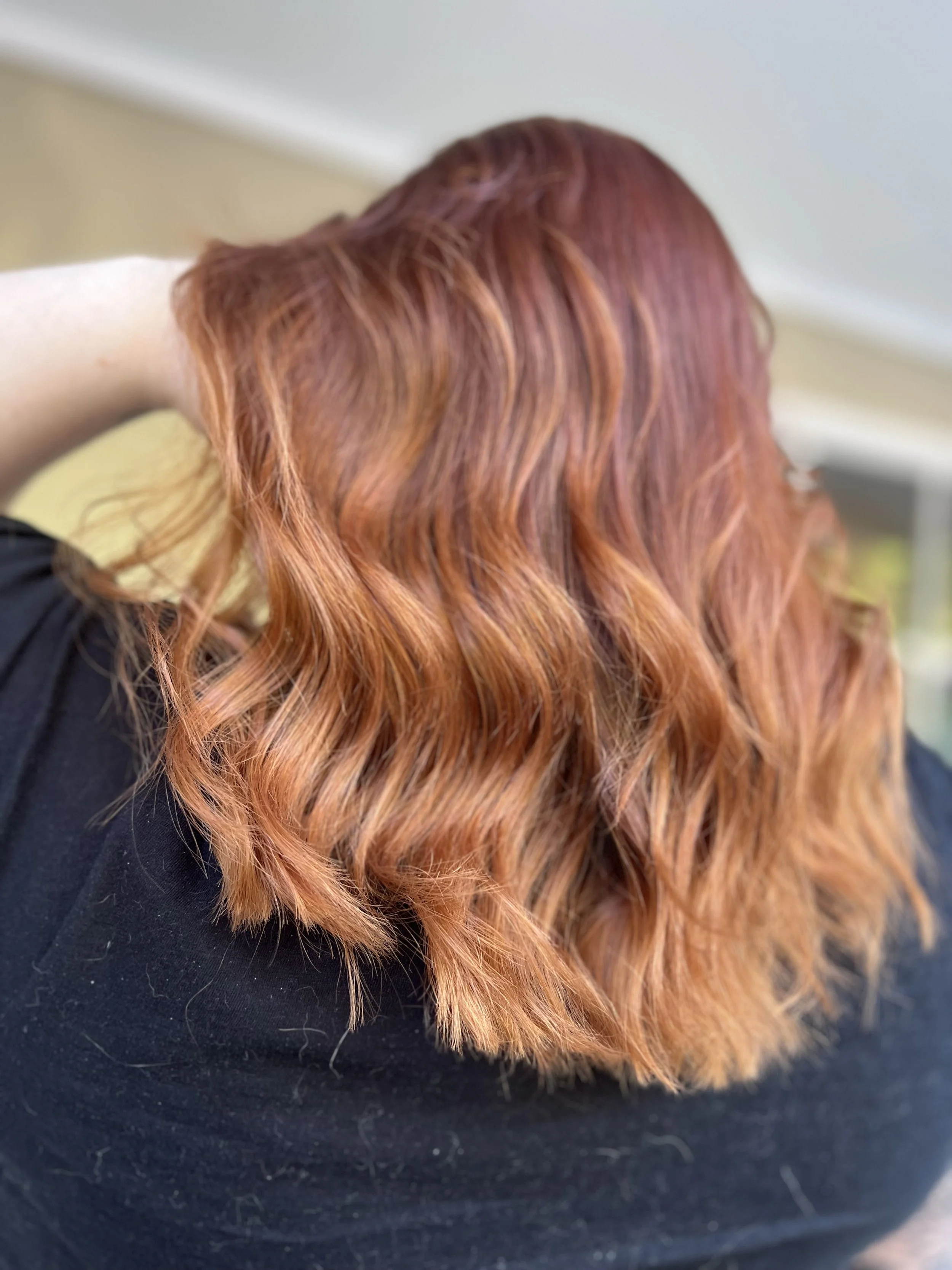 Close-up of a person's head showing shoulder-length, wavy red hair with subtle highlights, wearing a black shirt.