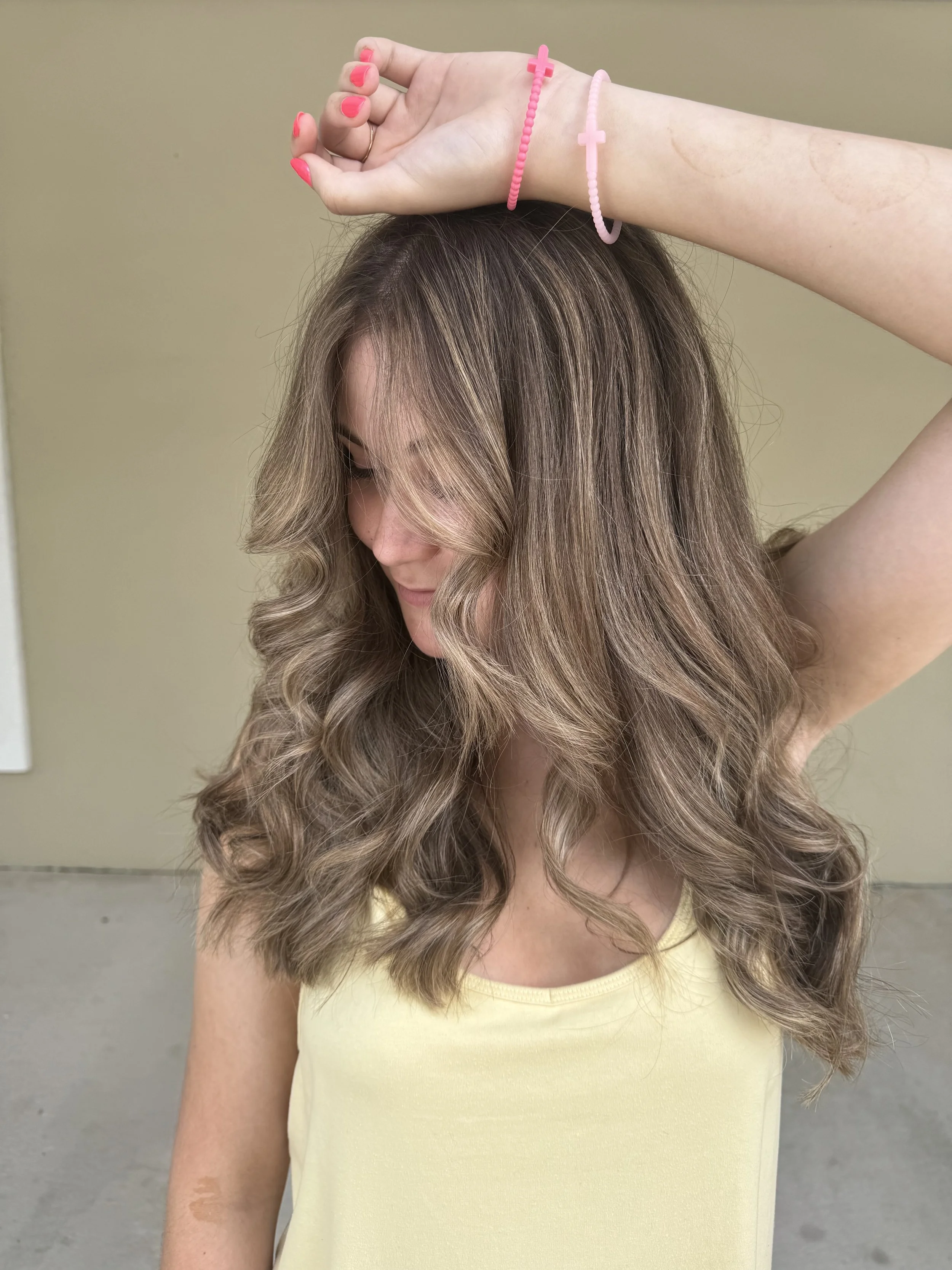 A young woman with wavy, light brown hair with blonde highlights, wearing a yellow sleeveless top, has one arm raised above her head. She is wearing pink bracelets with crosses on her wrist and her nails are painted pink. She is looking downward with