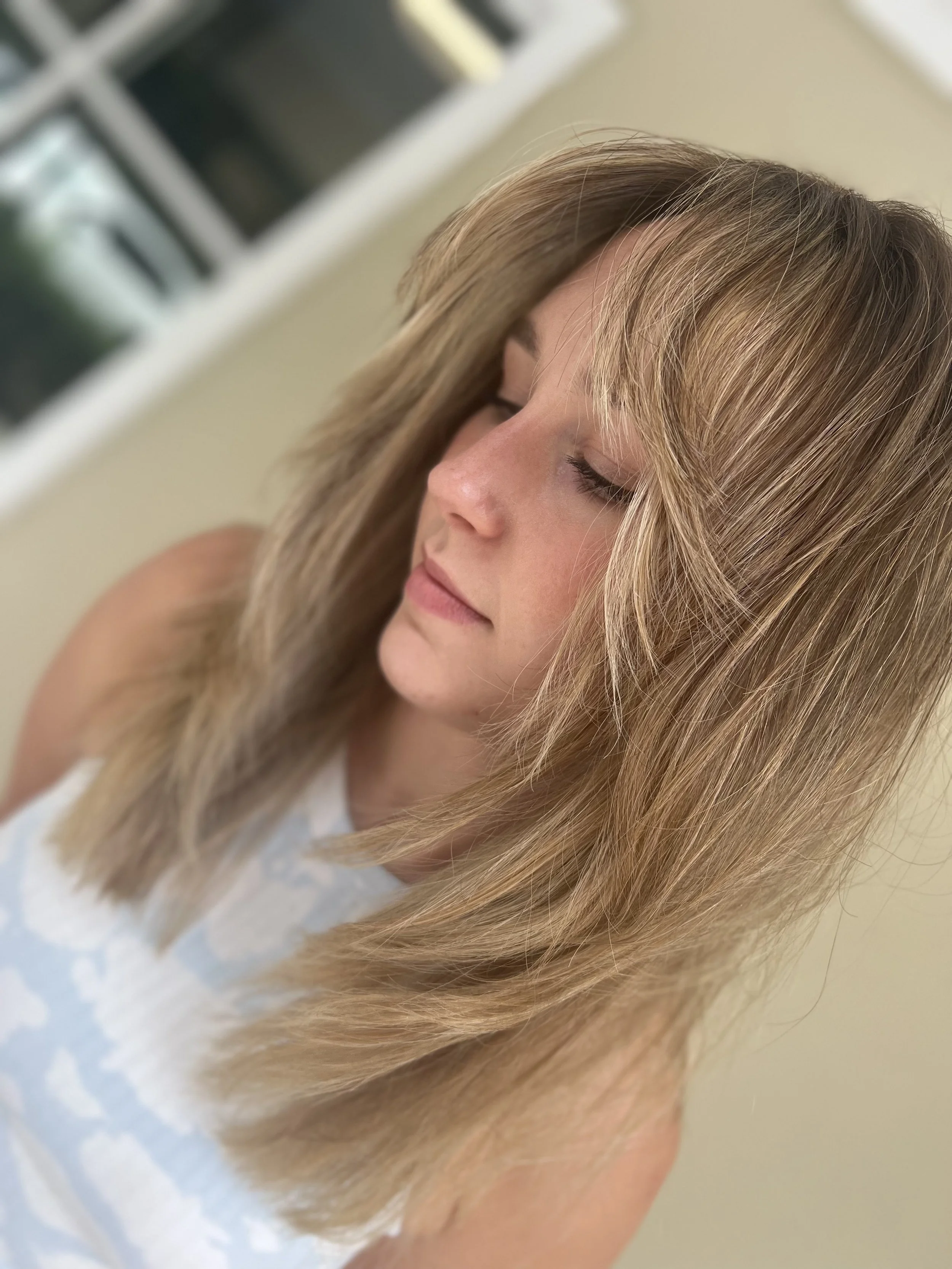 A woman with shoulder-length, layered blonde hair and side-swept bangs, with her eyes closed.