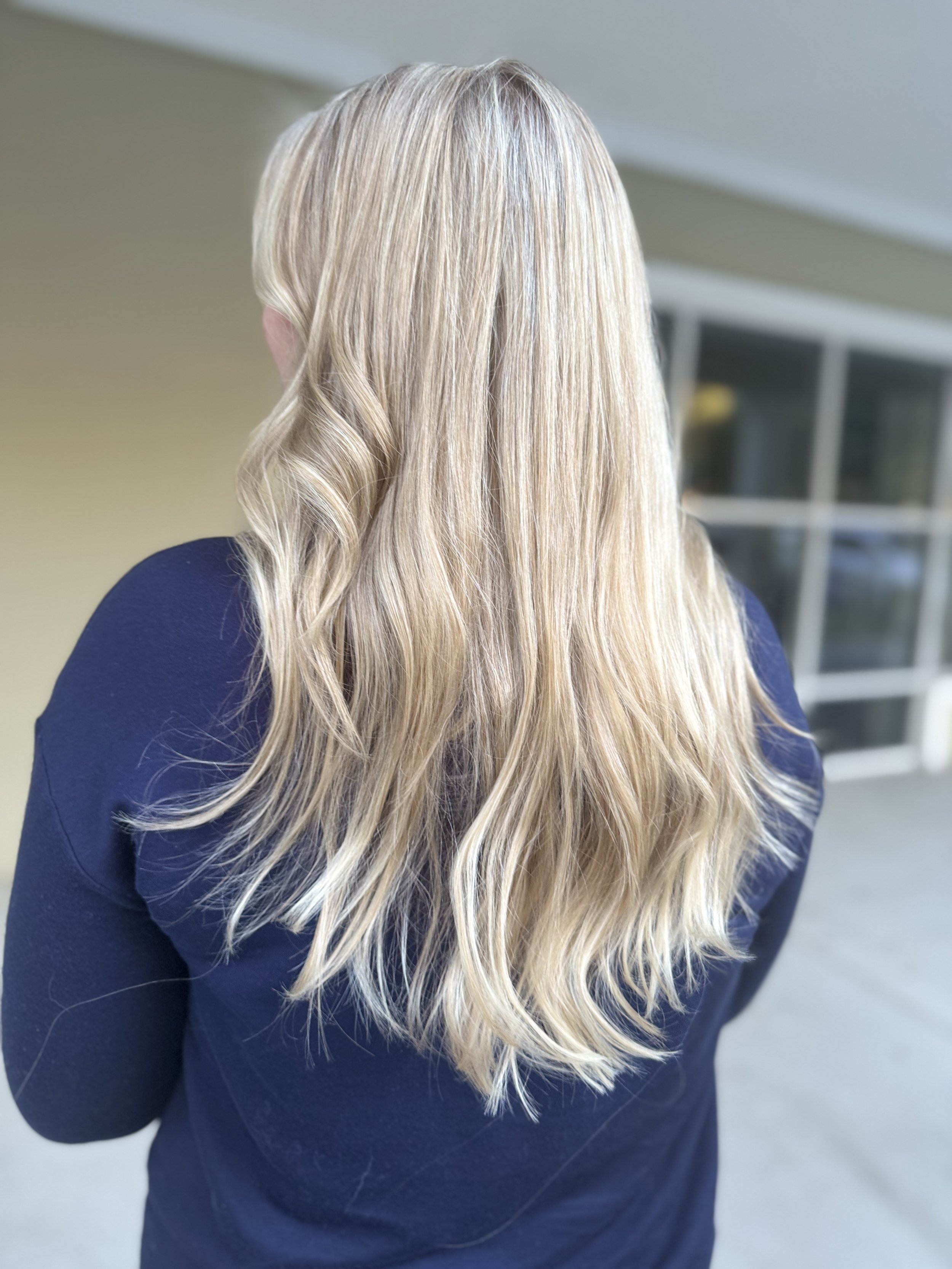 Back of a woman with long blonde wavy hair wearing a navy blue top, standing outside near a building.