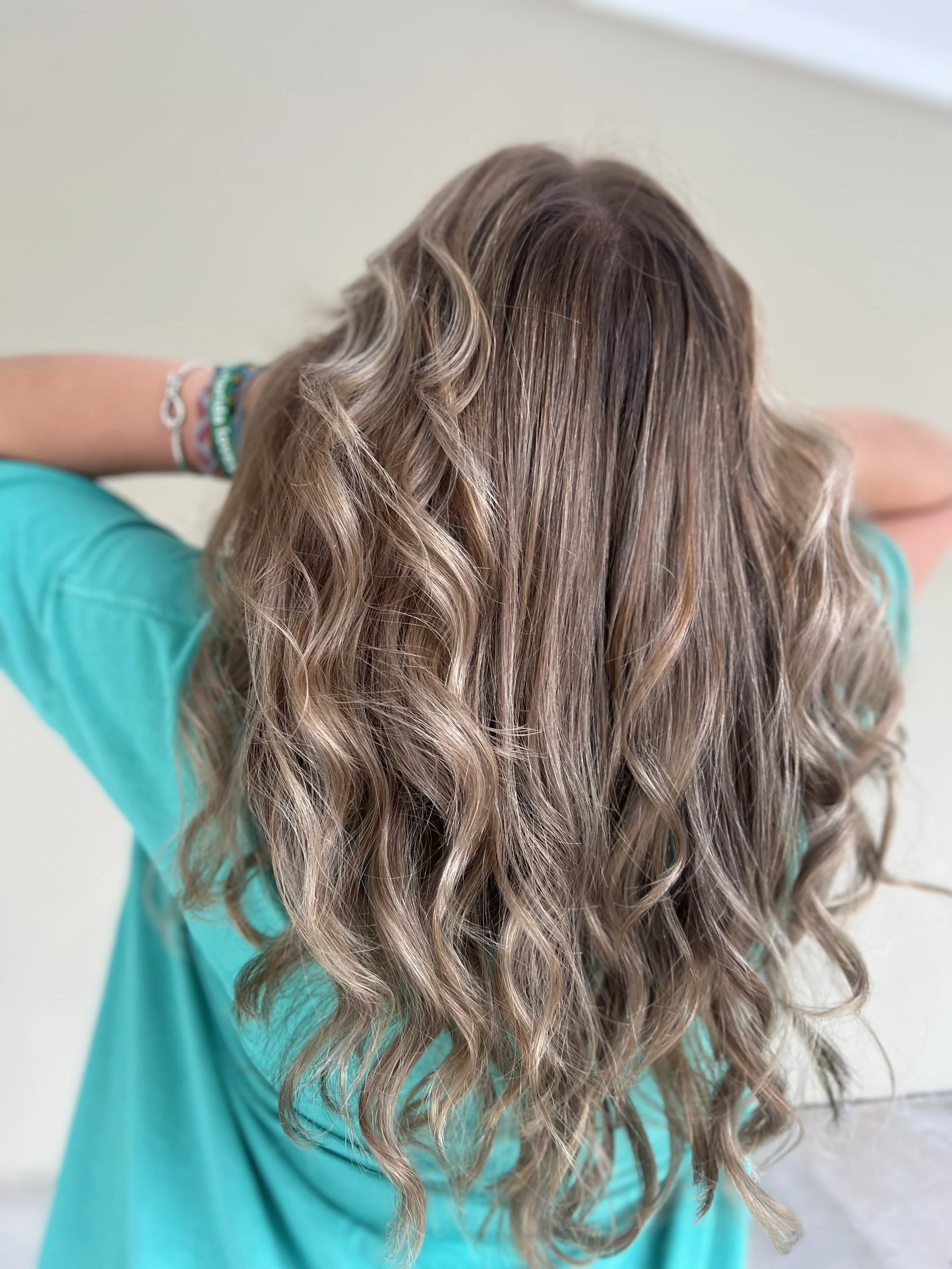 Back of a woman with long, wavy, light brown hair wearing a teal shirt and bracelets on her wrist.