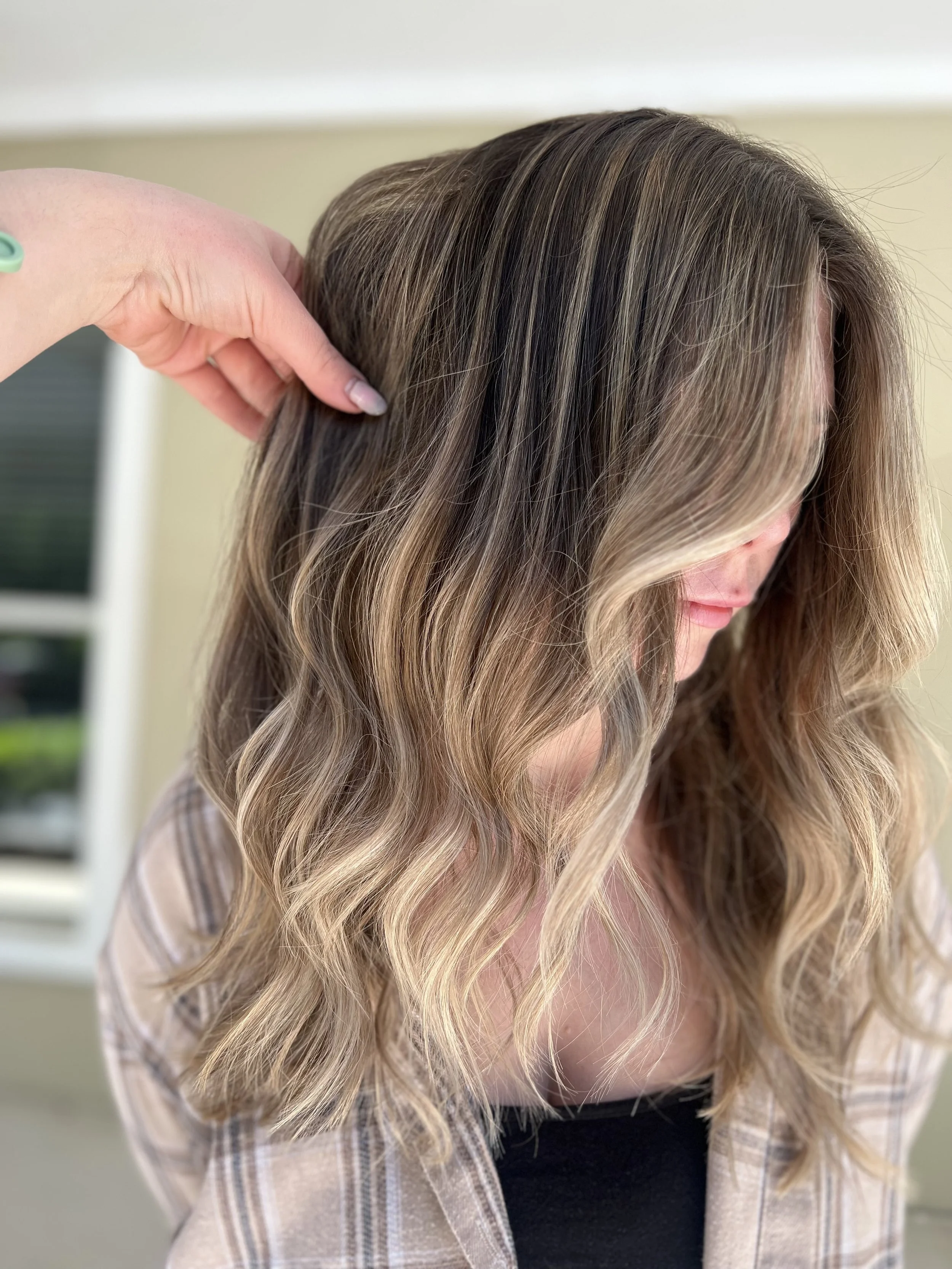 Close-up of a woman with wavy blonde hair and a plaid shirt, with a hand adjusting her hair.