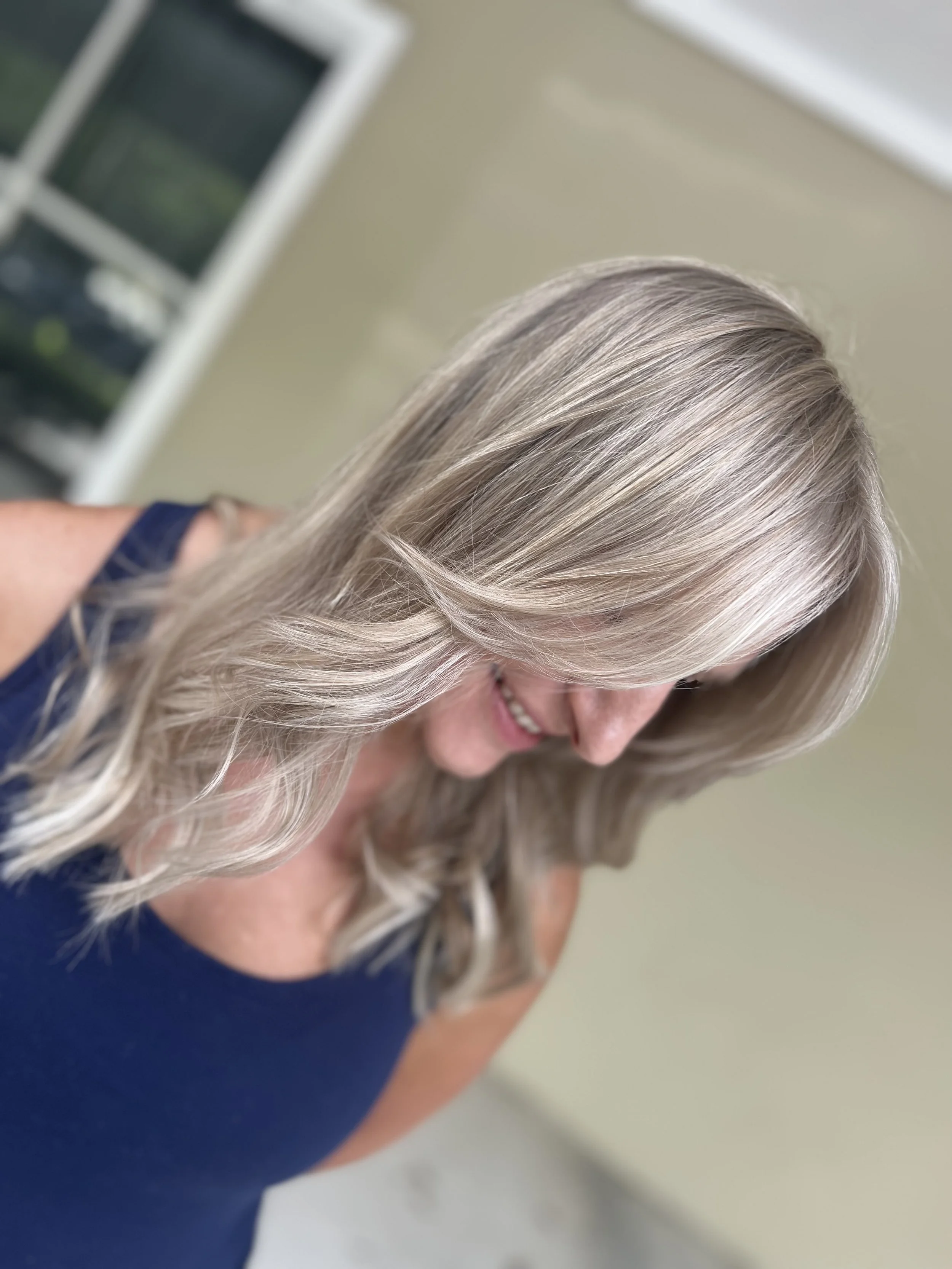 A woman with long, wavy, blonde hair smiling and looking downward.