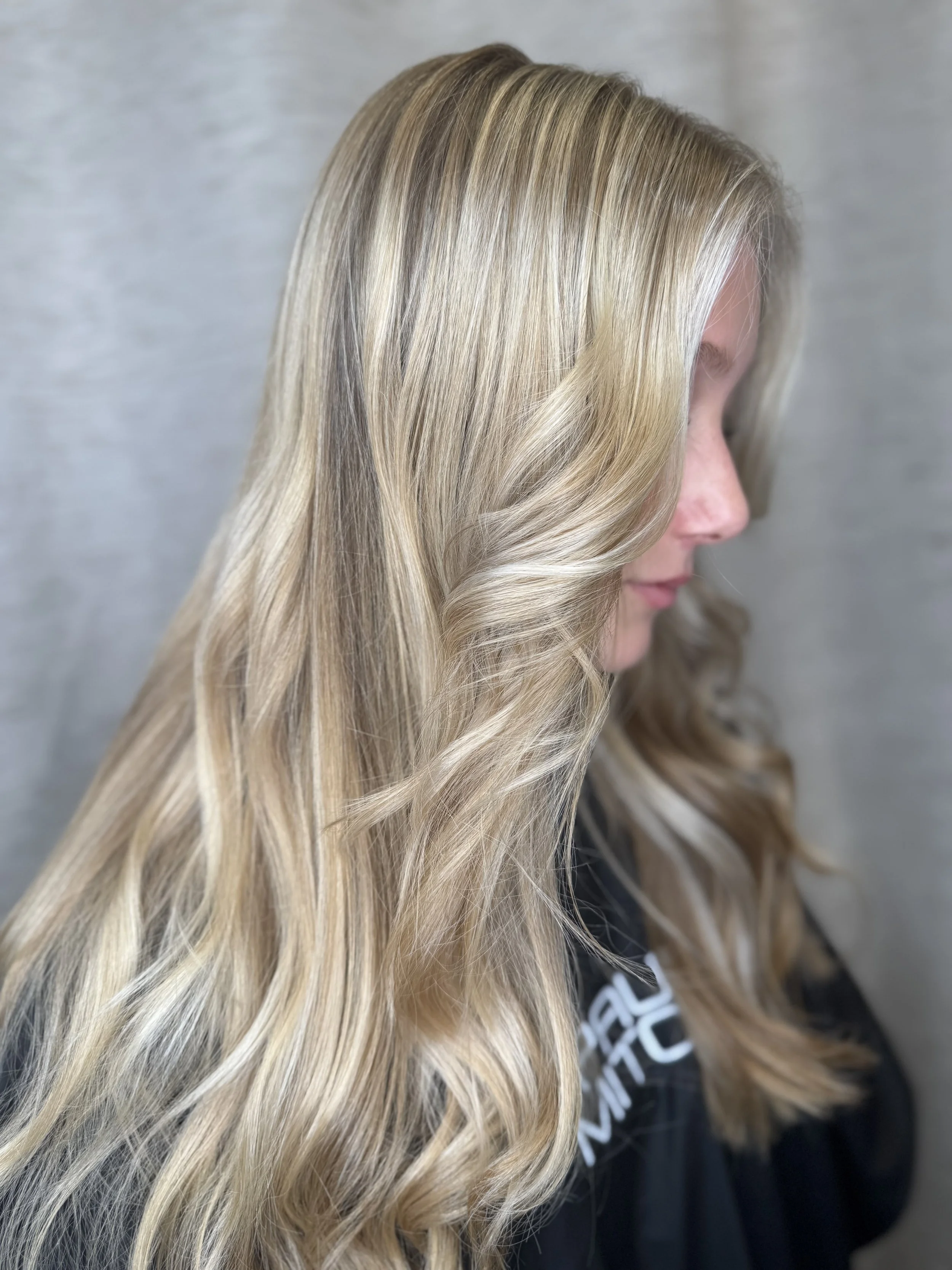 A woman with long, wavy blonde hair looking down, with a blurred gray background.