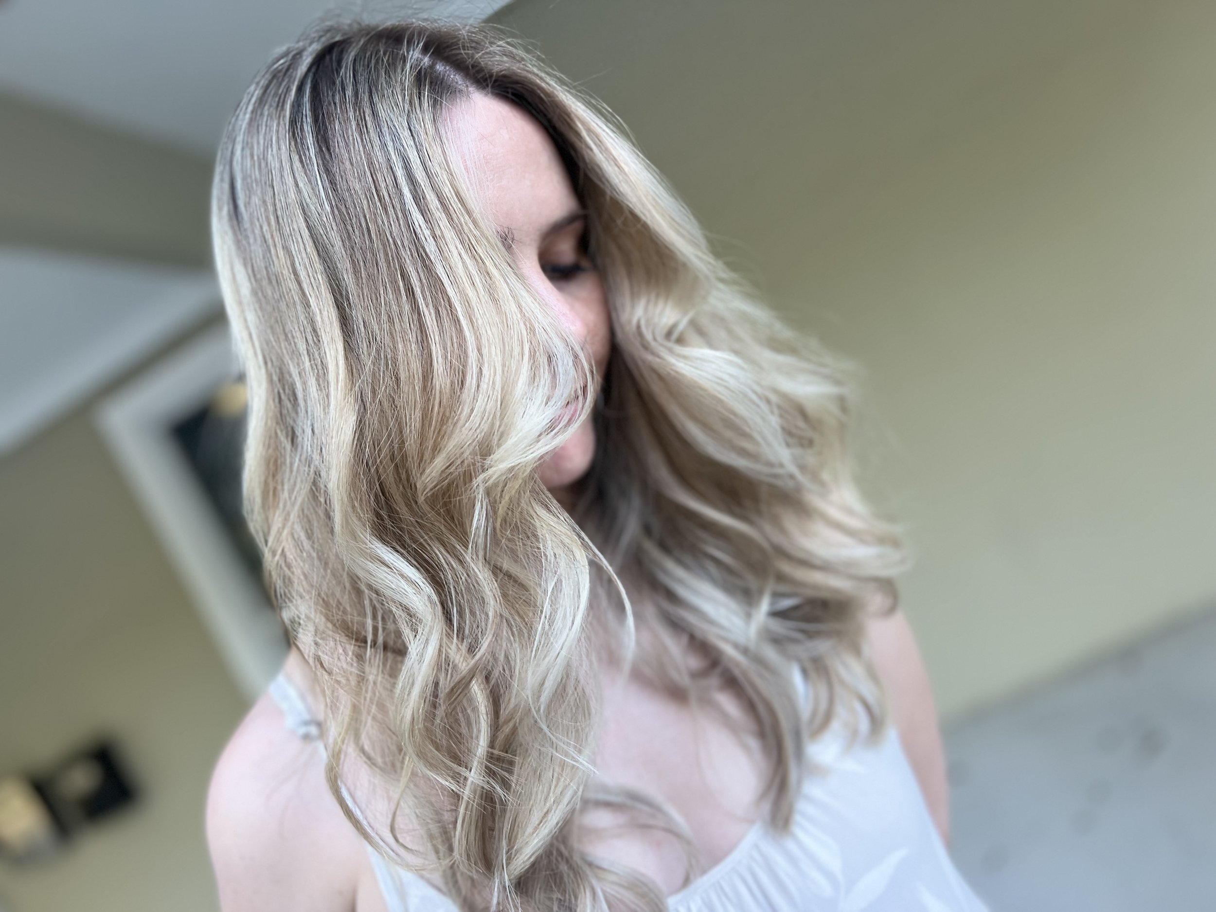 Close-up of a woman with long, wavy blonde hair looking down, wearing a white top, indoors with a blurred background.