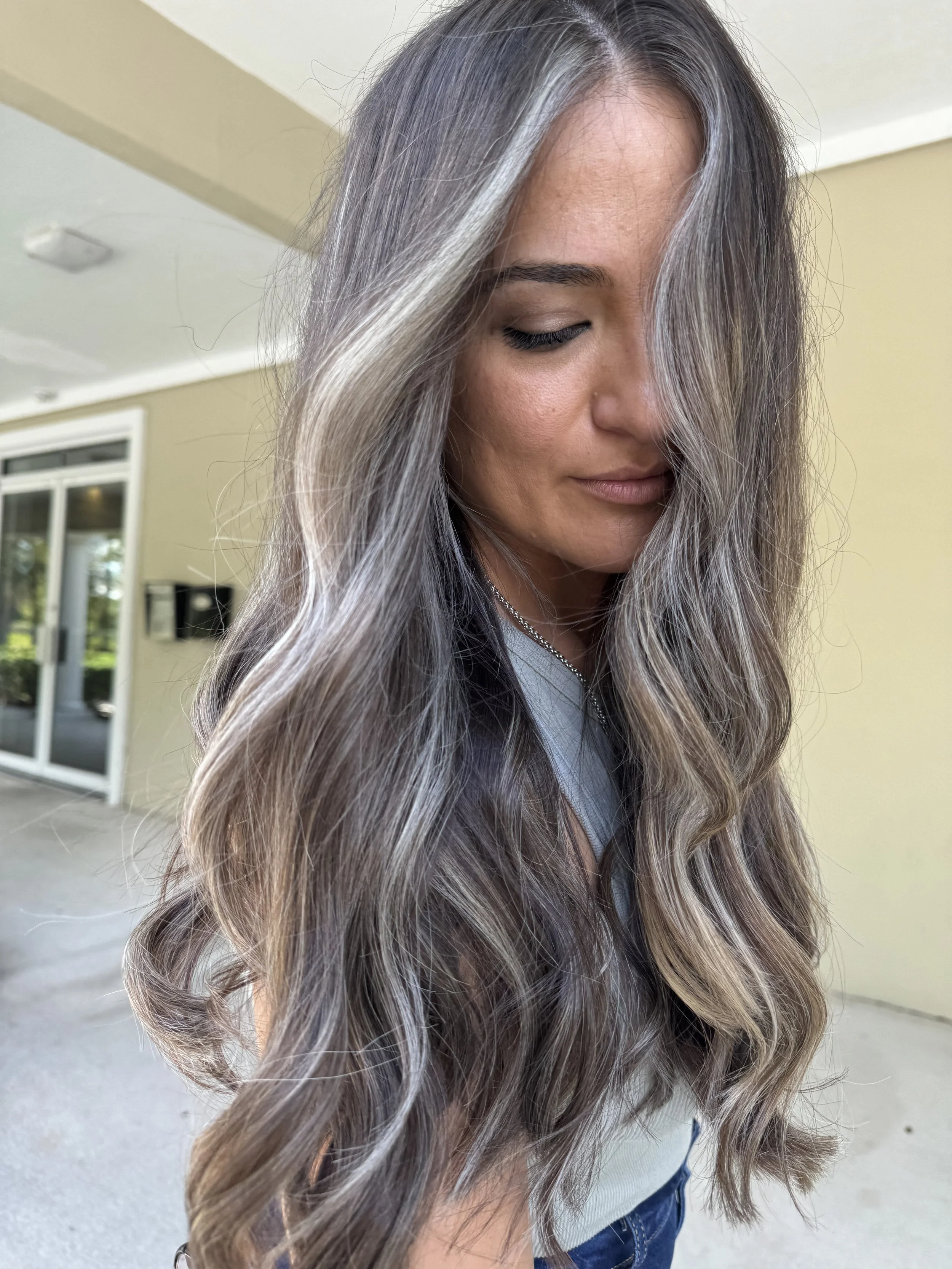 A woman with long, wavy gray hair looking down, standing outside near a beige wall and glass door.