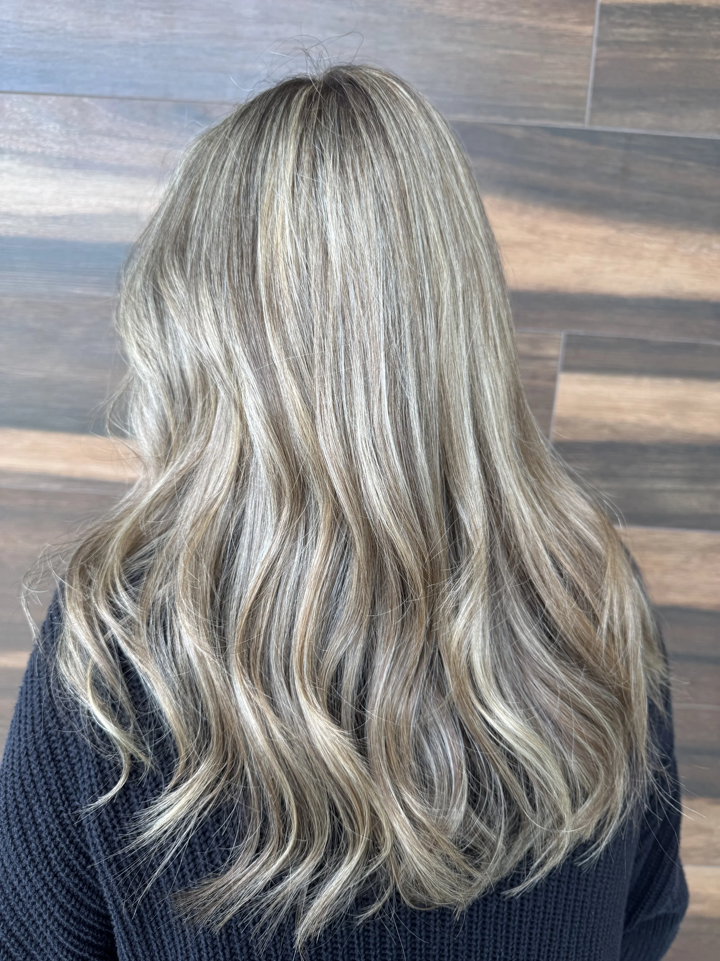 Back view of a woman with long, wavy blonde hair with highlights, wearing a dark sweater, standing in front of a wood-paneled wall.