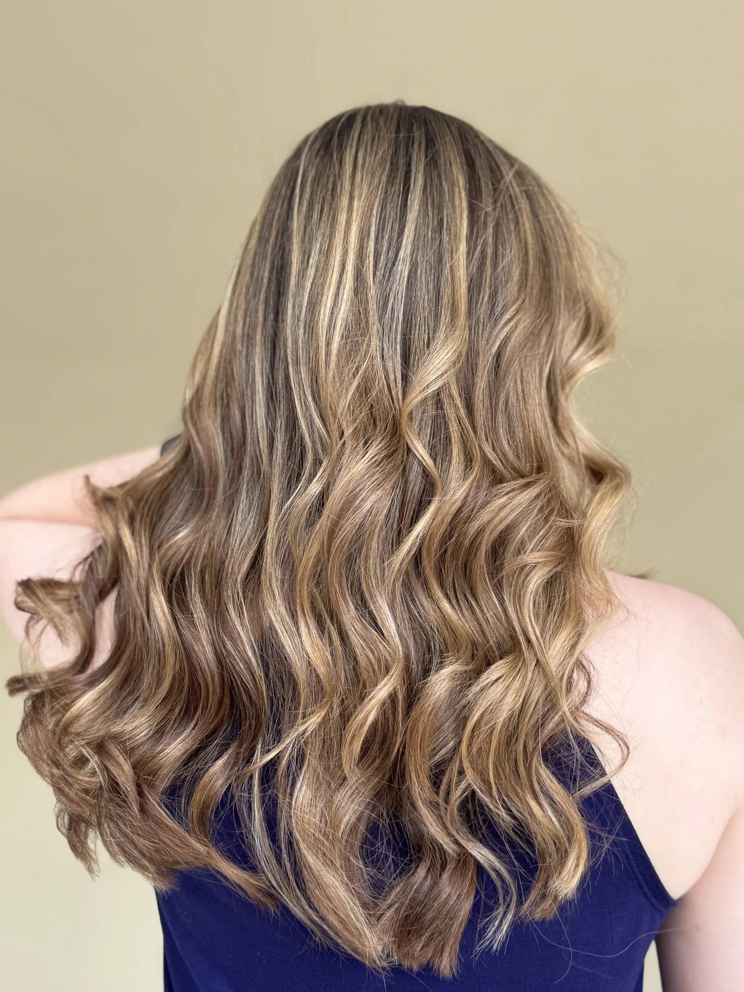 Woman with wavy, shoulder-length light brown hair with blonde highlights, wearing a navy blue top, facing away from the camera.