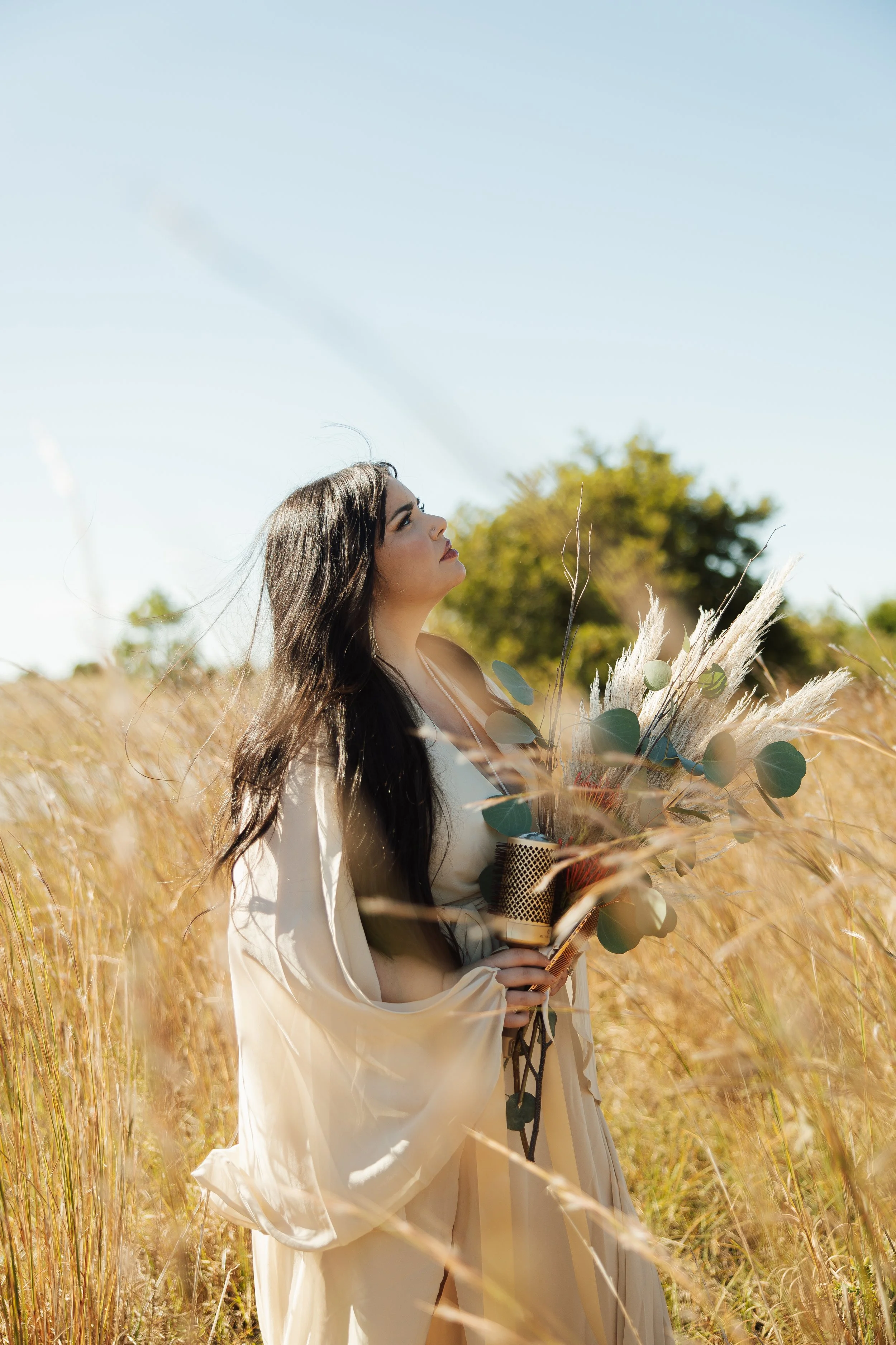 A woman with long dark hair standing in a grassy field, holding a bouquet of dried flowers and pampas grass, looking up at the sky on a sunny day.