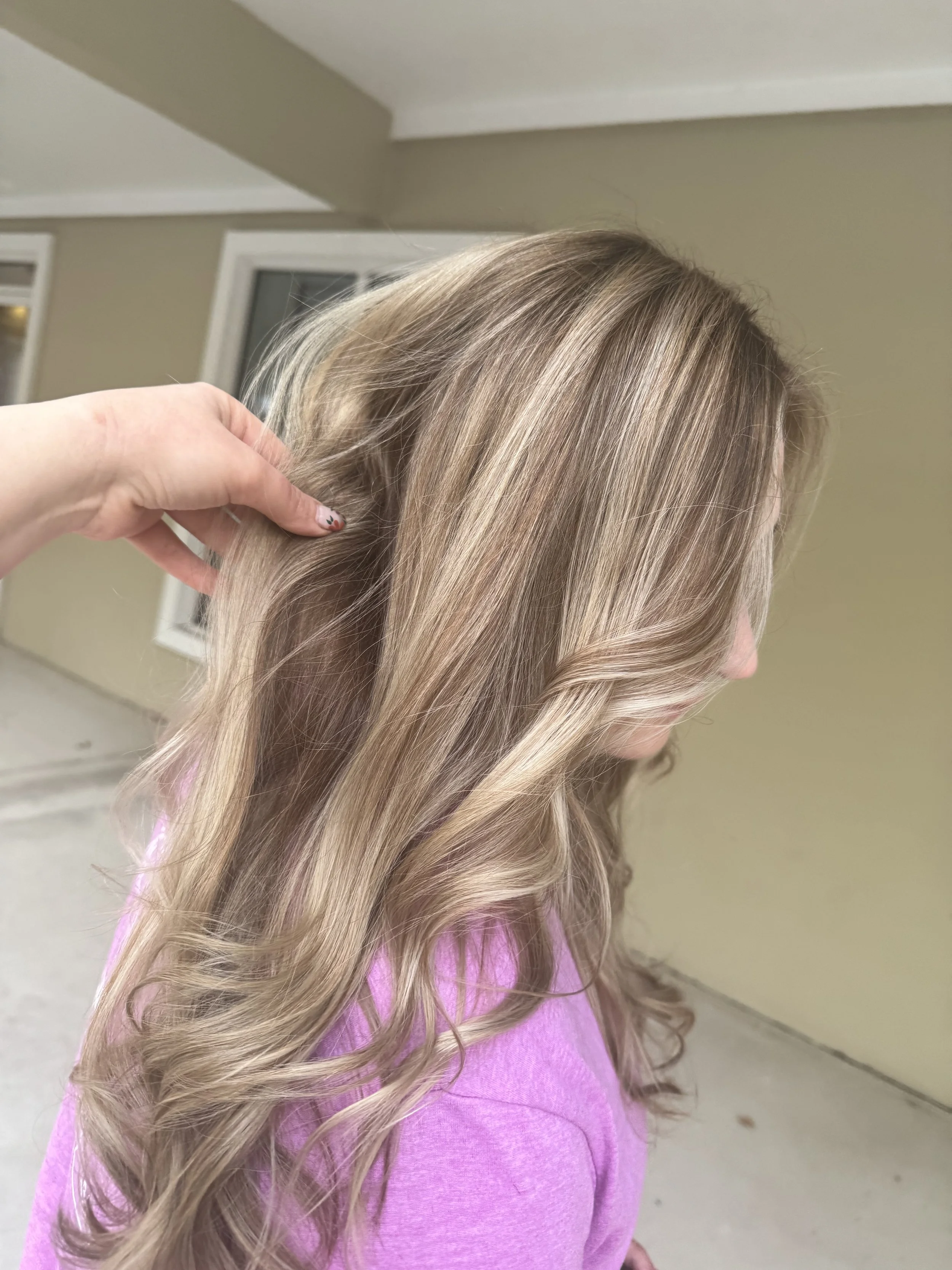 A person with long, wavy blonde hair wearing a pink shirt. Someone's hand is holding a section of their hair, showing its color and texture.