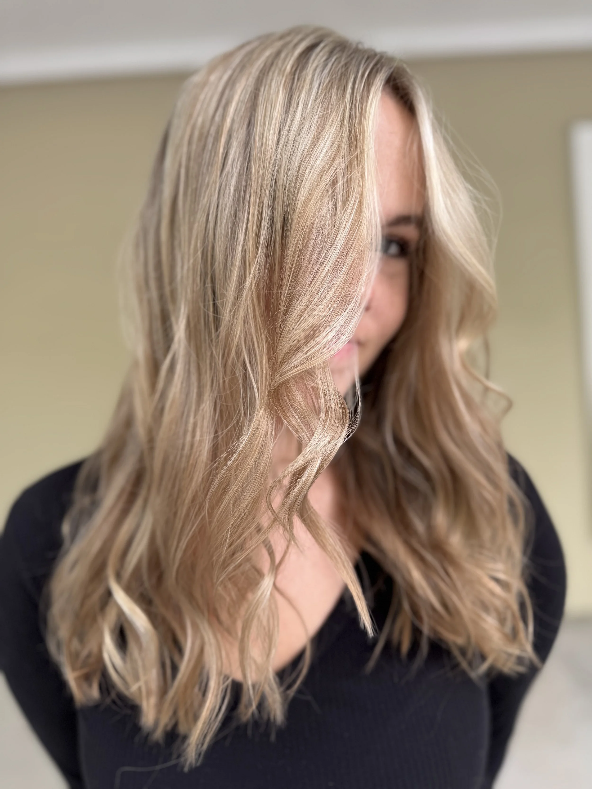 A woman with wavy blonde hair in a loose style, wearing a black top, looking to the side with a blurred indoor background.