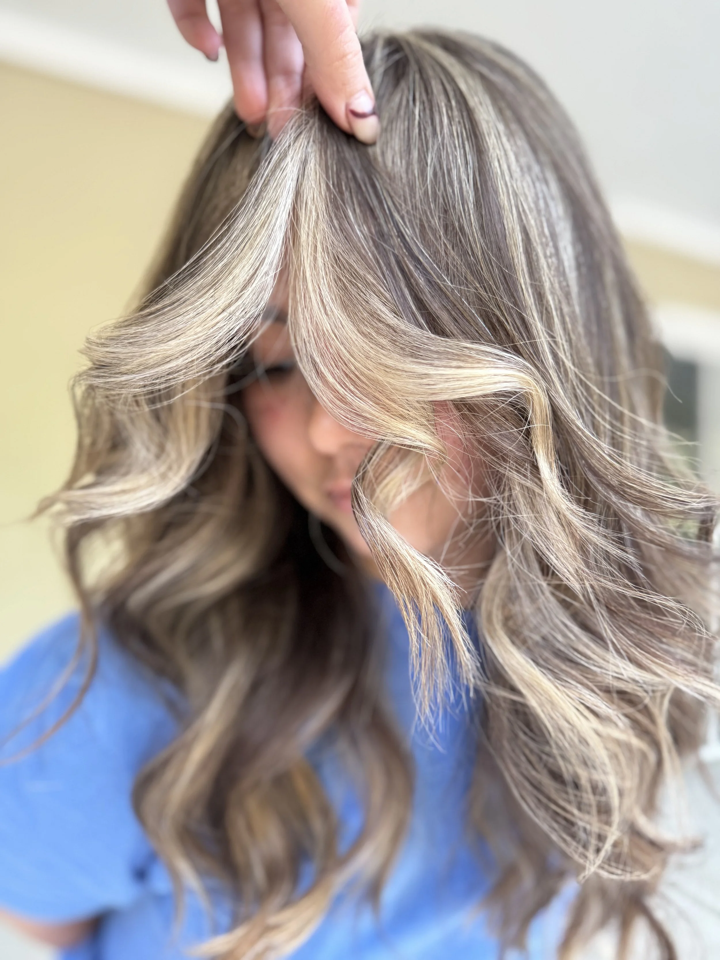 A woman with long, wavy, blonde hair, styled in loose curls, is having her hair touched or adjusted by a hand.