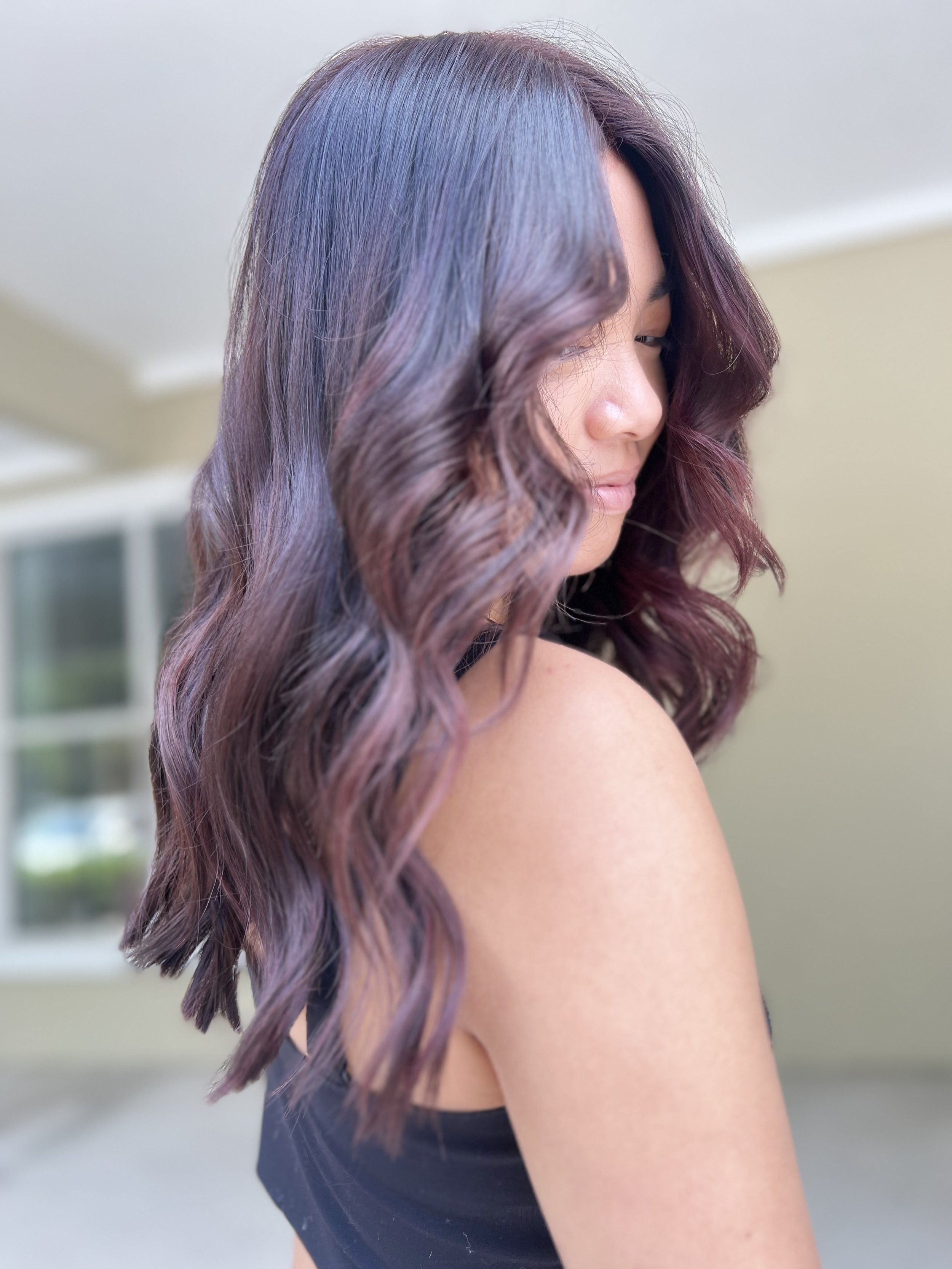 Close-up of a woman with wavy, shoulder-length, multi-colored hair including shades of purple and blue, wearing a black top, indoors with a blurred background.