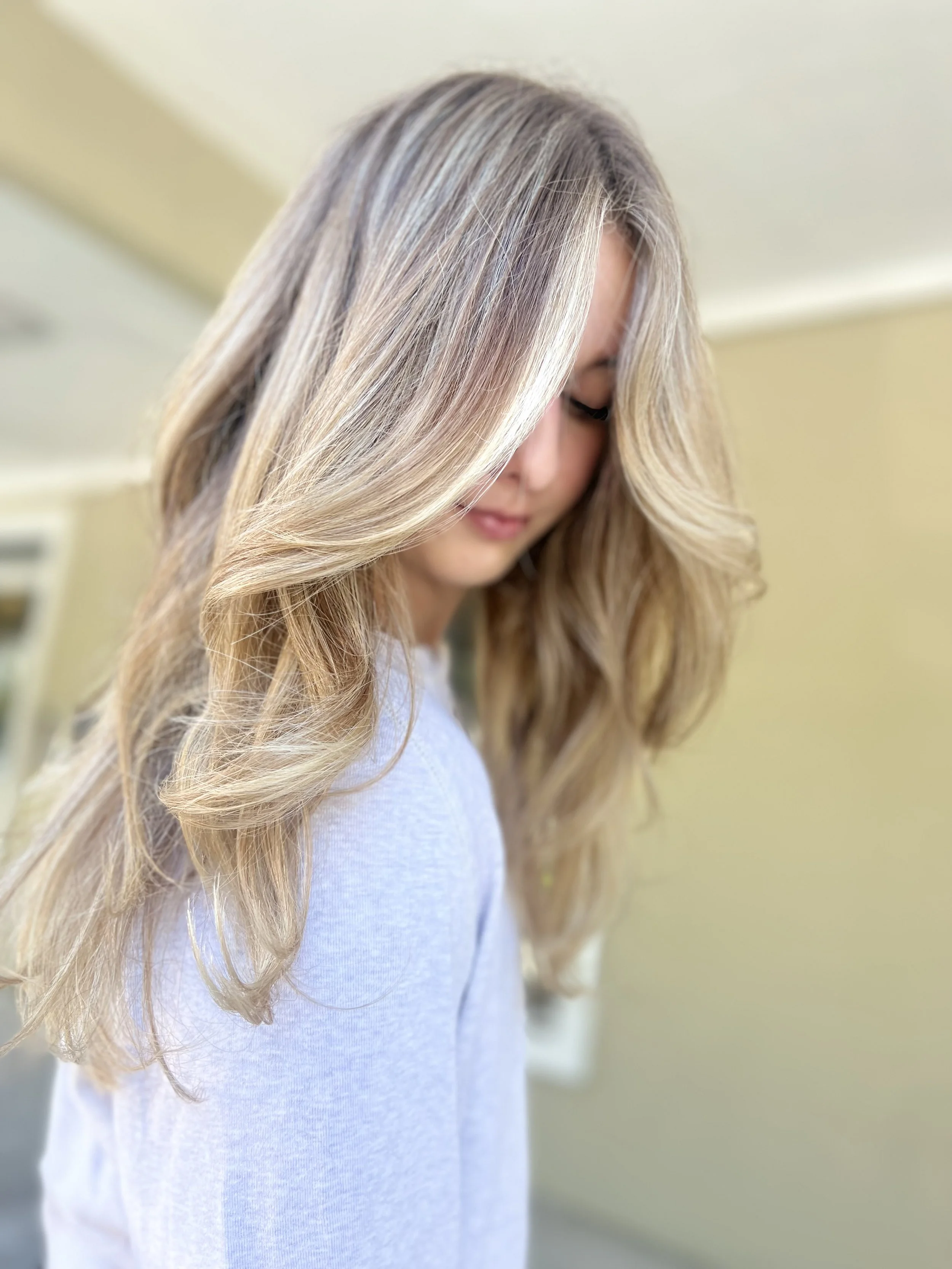 Close-up of a woman with long, wavy blonde hair, wearing a light gray top, looking down with her eyes closed.