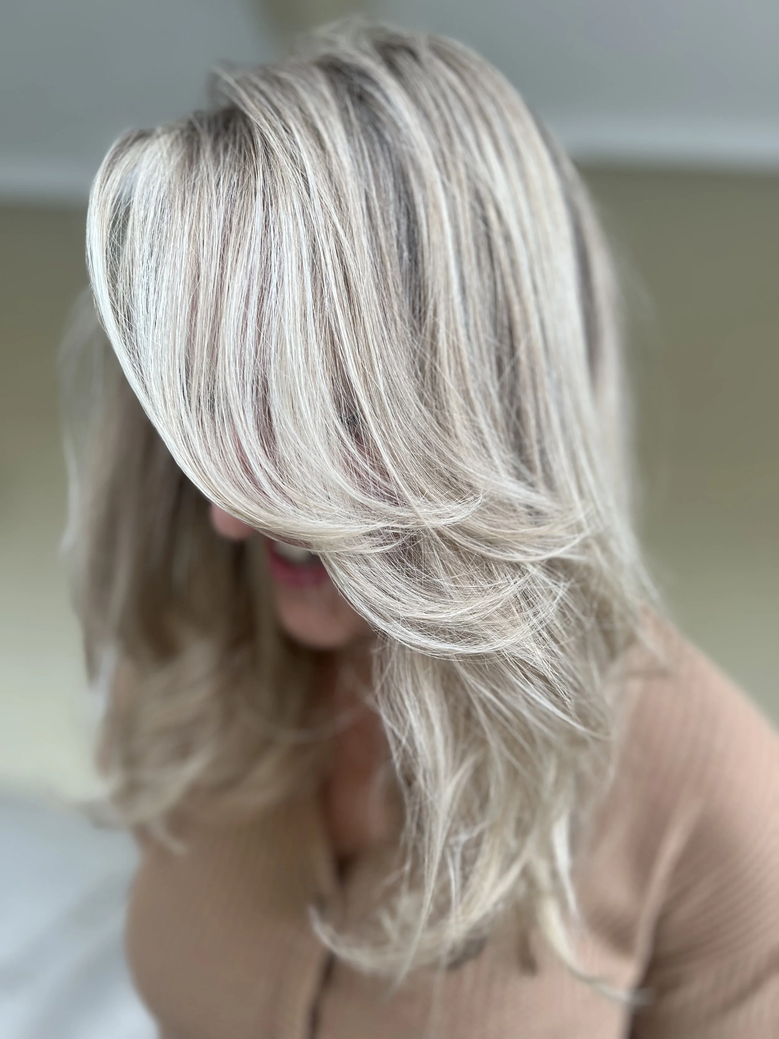Close-up of a woman with long, layered, blonde hair with highlights, wearing a beige top, with her face partially obscured by her hair.