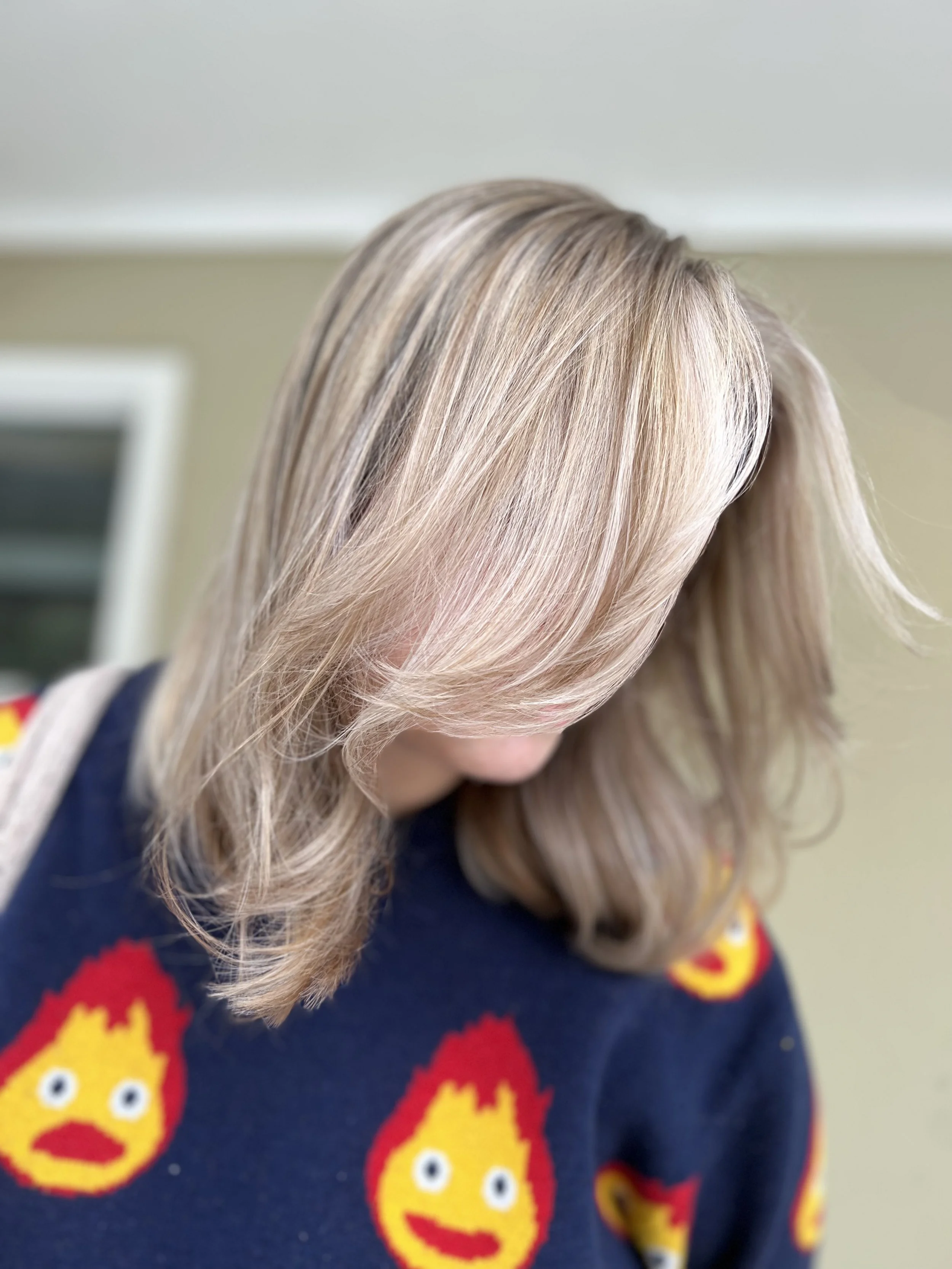 Close-up of a woman with shoulder-length blonde hair, looking downward, wearing a dark sweater with colorful flame and face patterns.