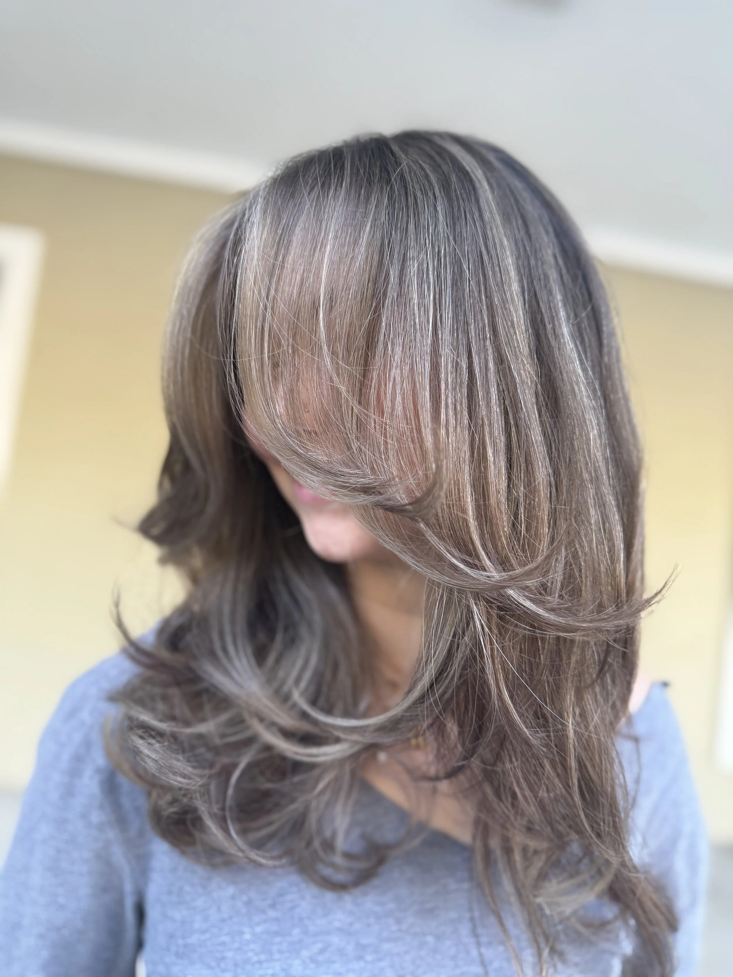 Woman with multi-shaded brown hair styled in loose waves, wearing a gray top, looking down.