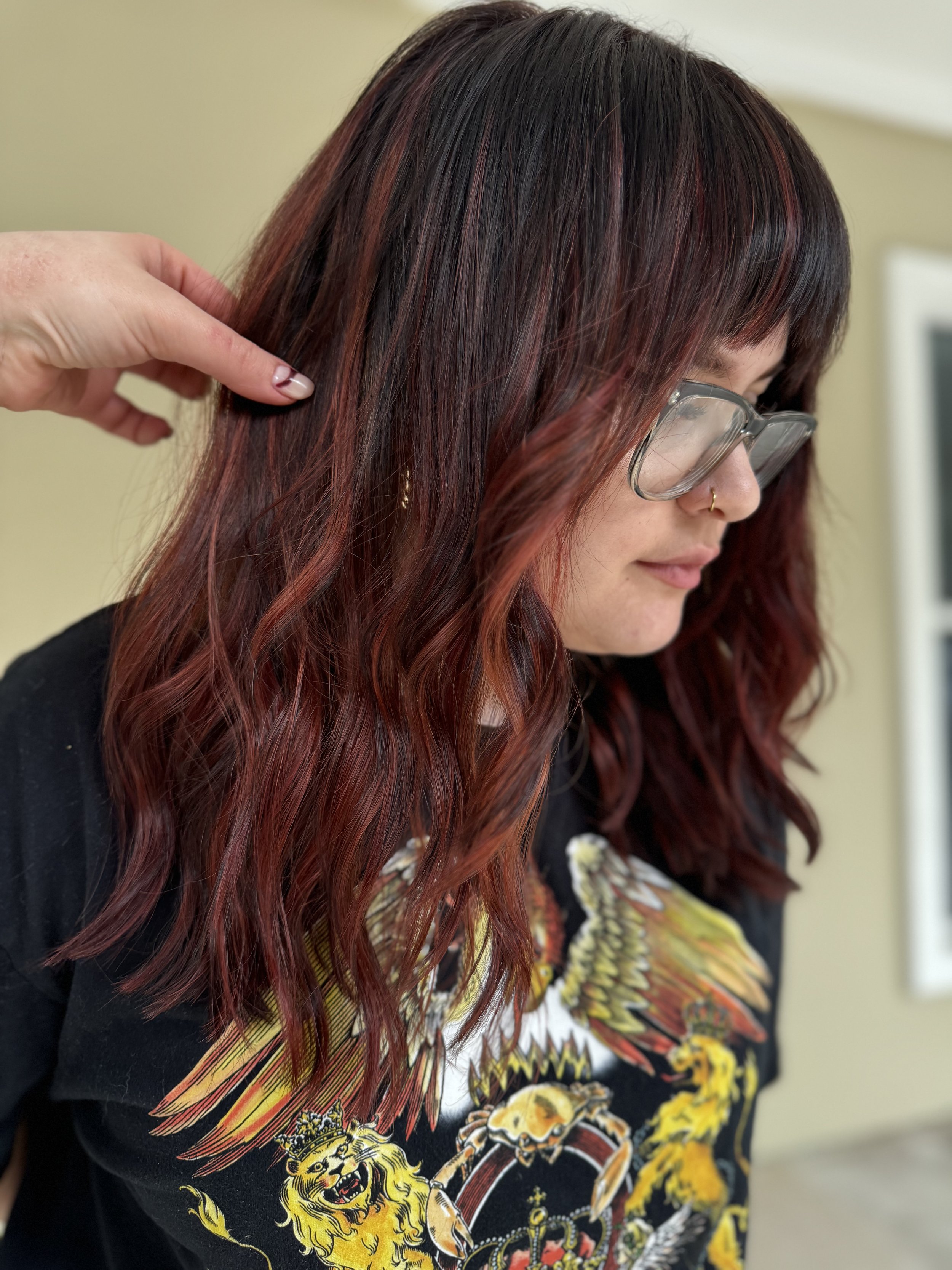A woman with wavy, burgundy hair, wearing glasses and a graphic T-shirt, is getting her hair styled by someone off-camera.