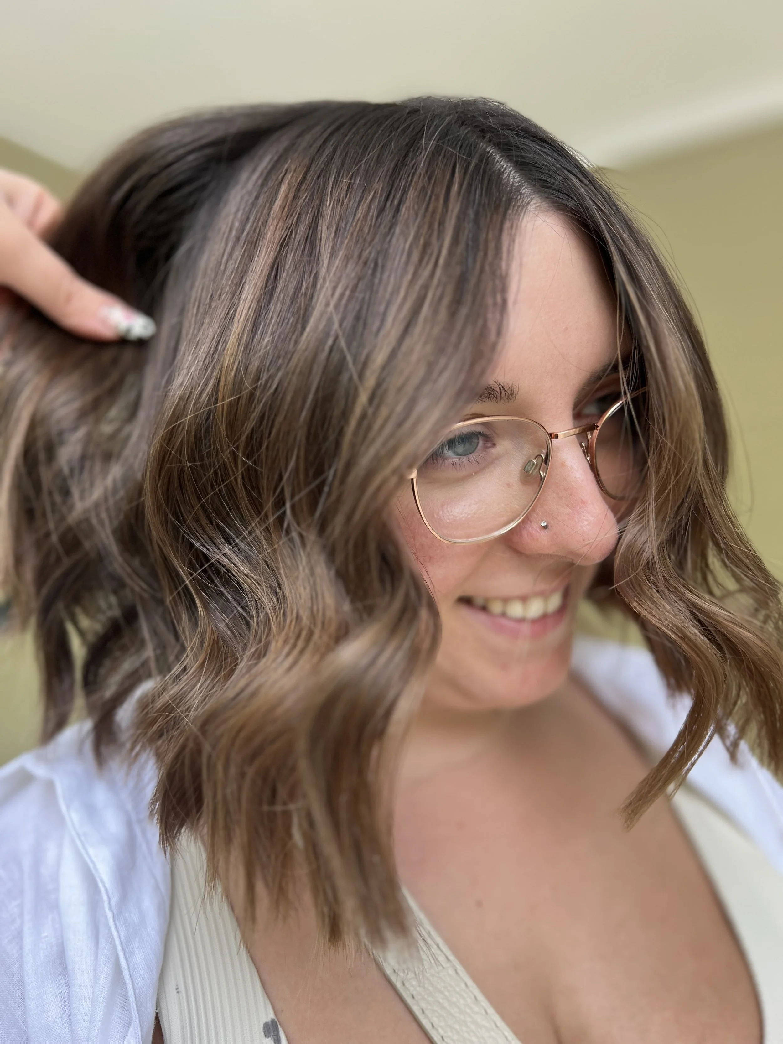 Close-up of a woman with shoulder-length wavy brown hair and glasses, smiling and looking down, with a light background.