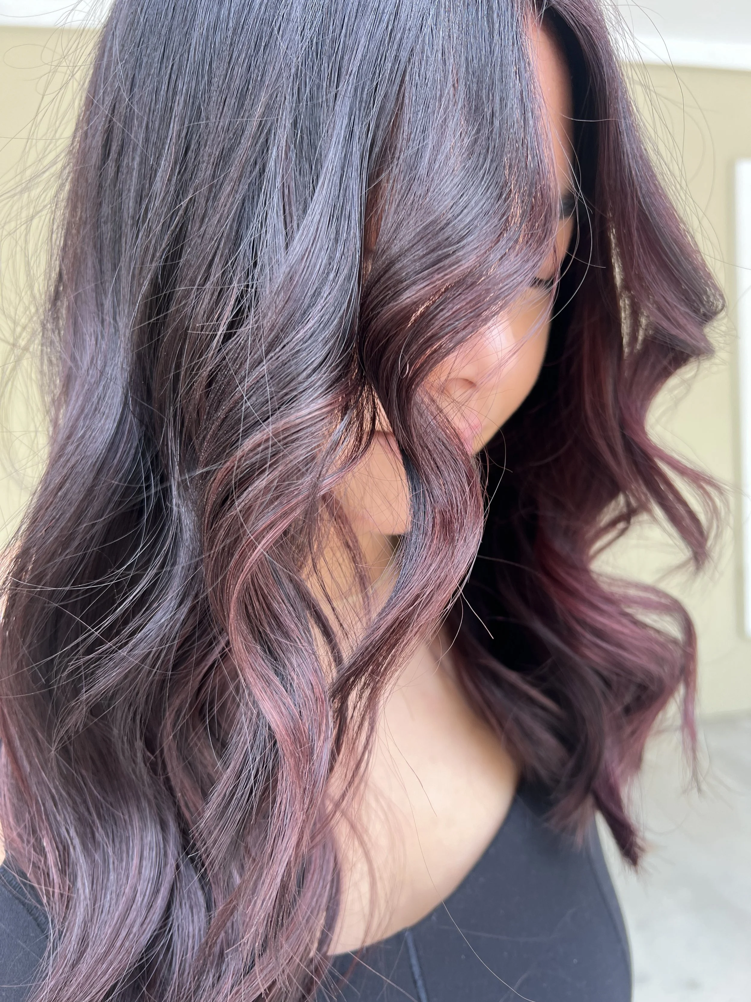 Close-up of a woman's wavy hair with black and purple hues, partially covering her face, with a blurred indoor background.