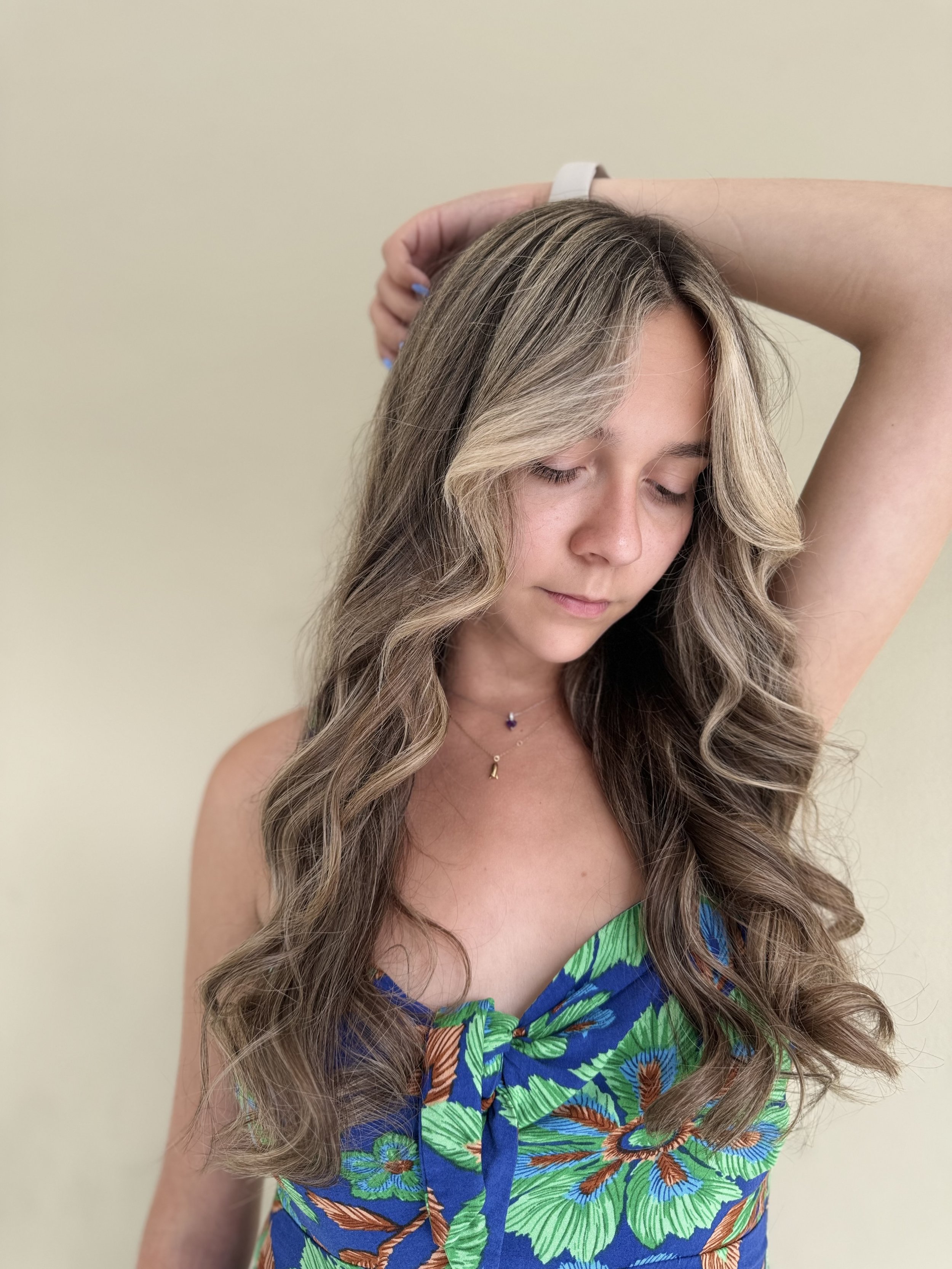 A young woman with wavy, blonde hair with darker roots, wearing a strapless tropical dress with green and blue leaves and flowers, and a delicate necklace with a small purple and gold pendant, posing with one arm raised and her eyes closed against a 